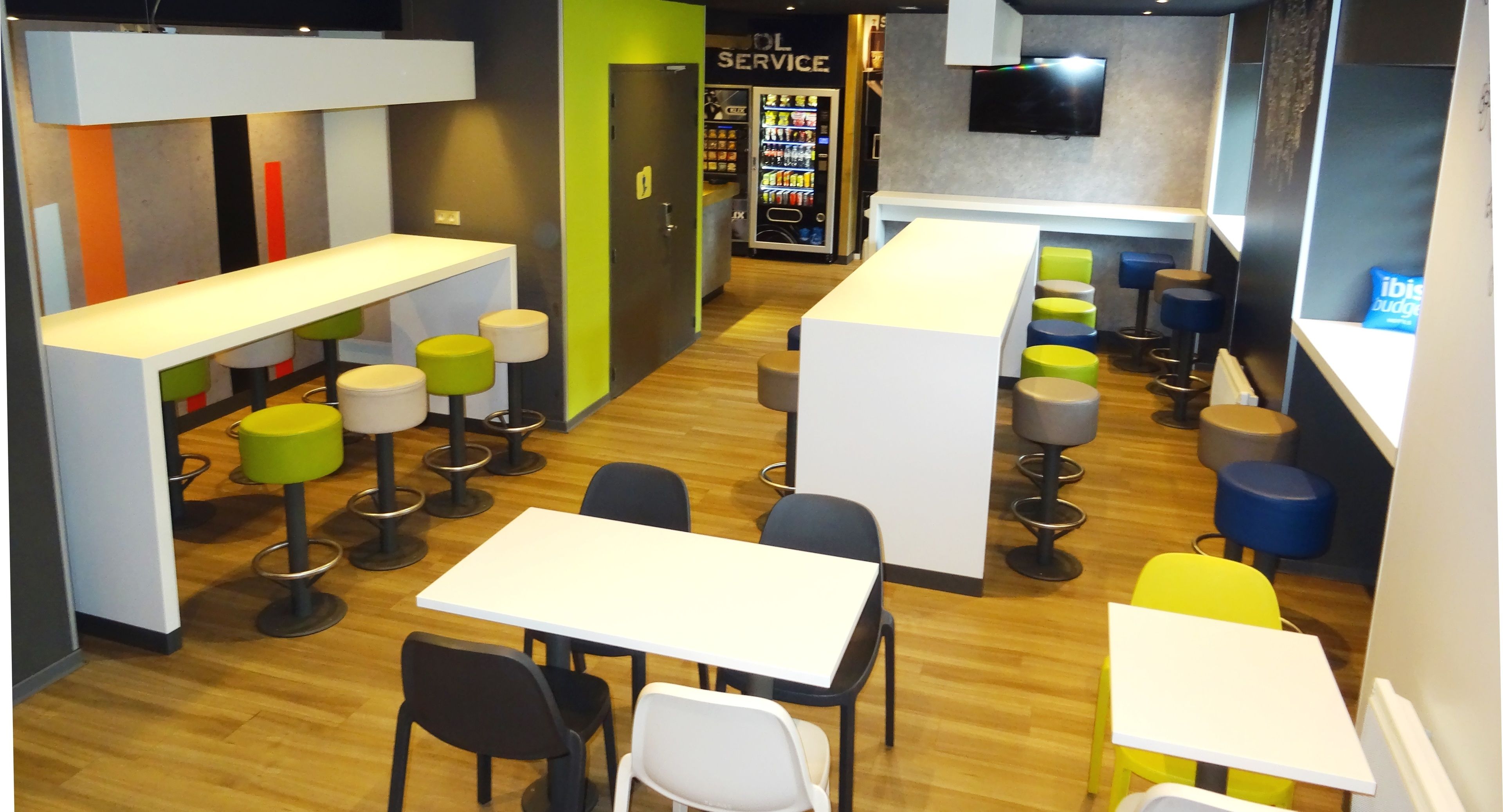 A modern bar area with white tables and colorful stools, a TV mounted on the wall, and a vending machine in the background.