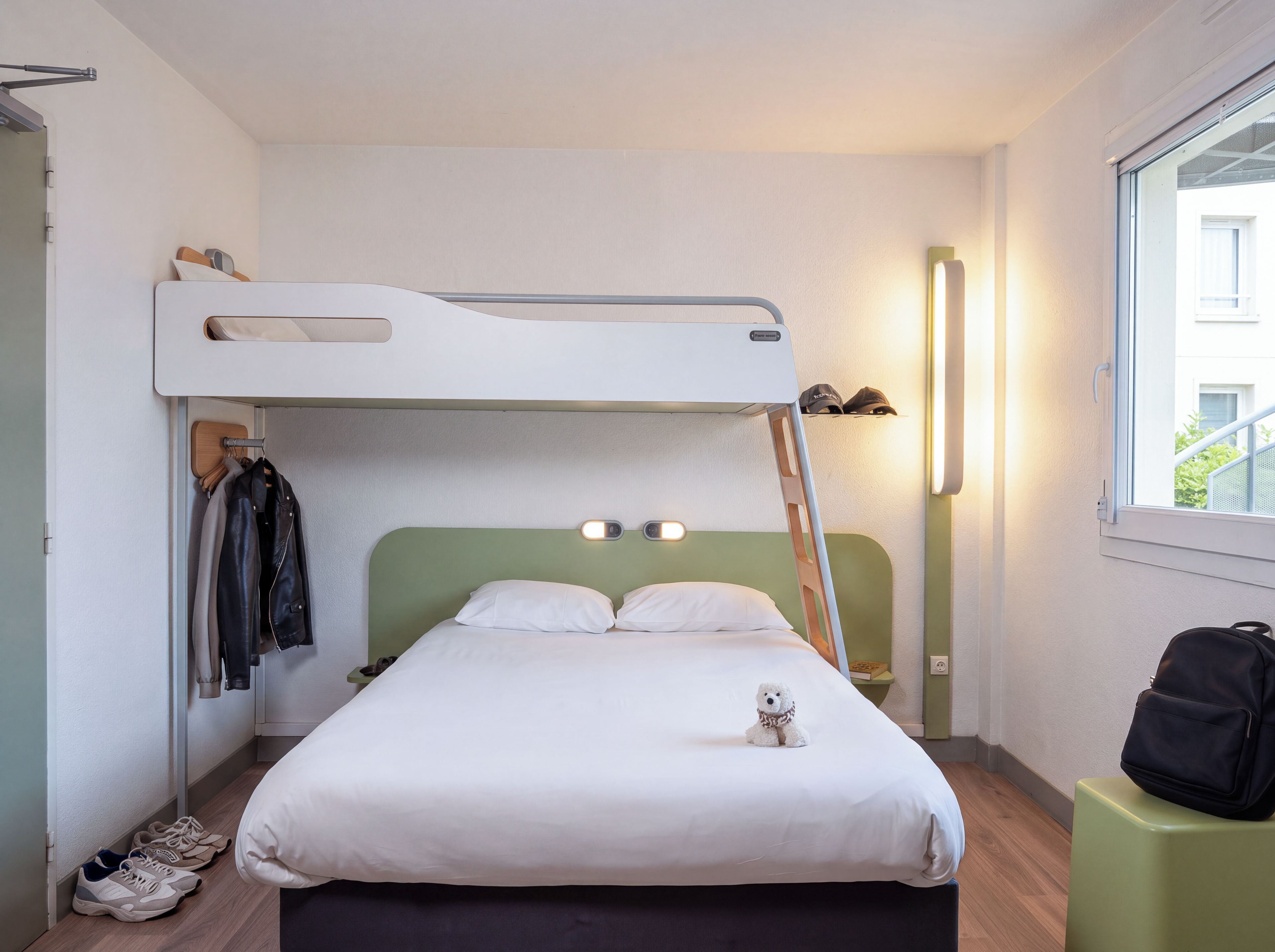 A bunk bed with a desk and chair, a small table with a lamp, and a window with a view of the outside.