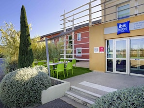 An outdoor seating area with green chairs and tables, a hedge, and a building with an 'ibis budget' sign.