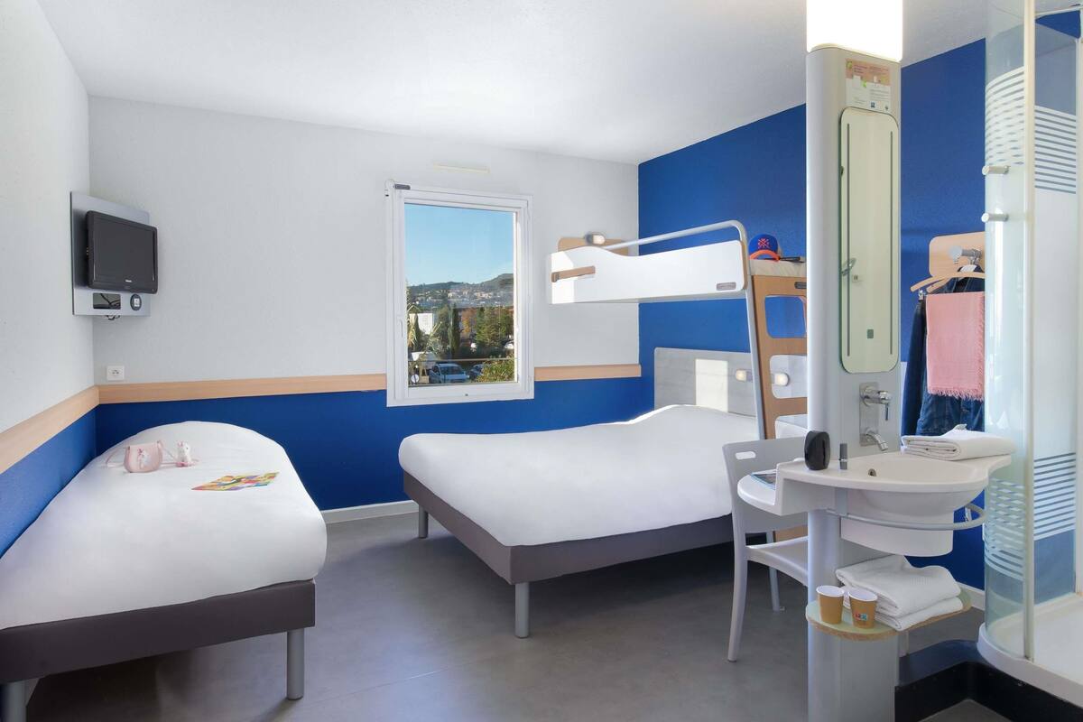 A hotel room with two beds, a desk, a mirror, a sink, and a TV.