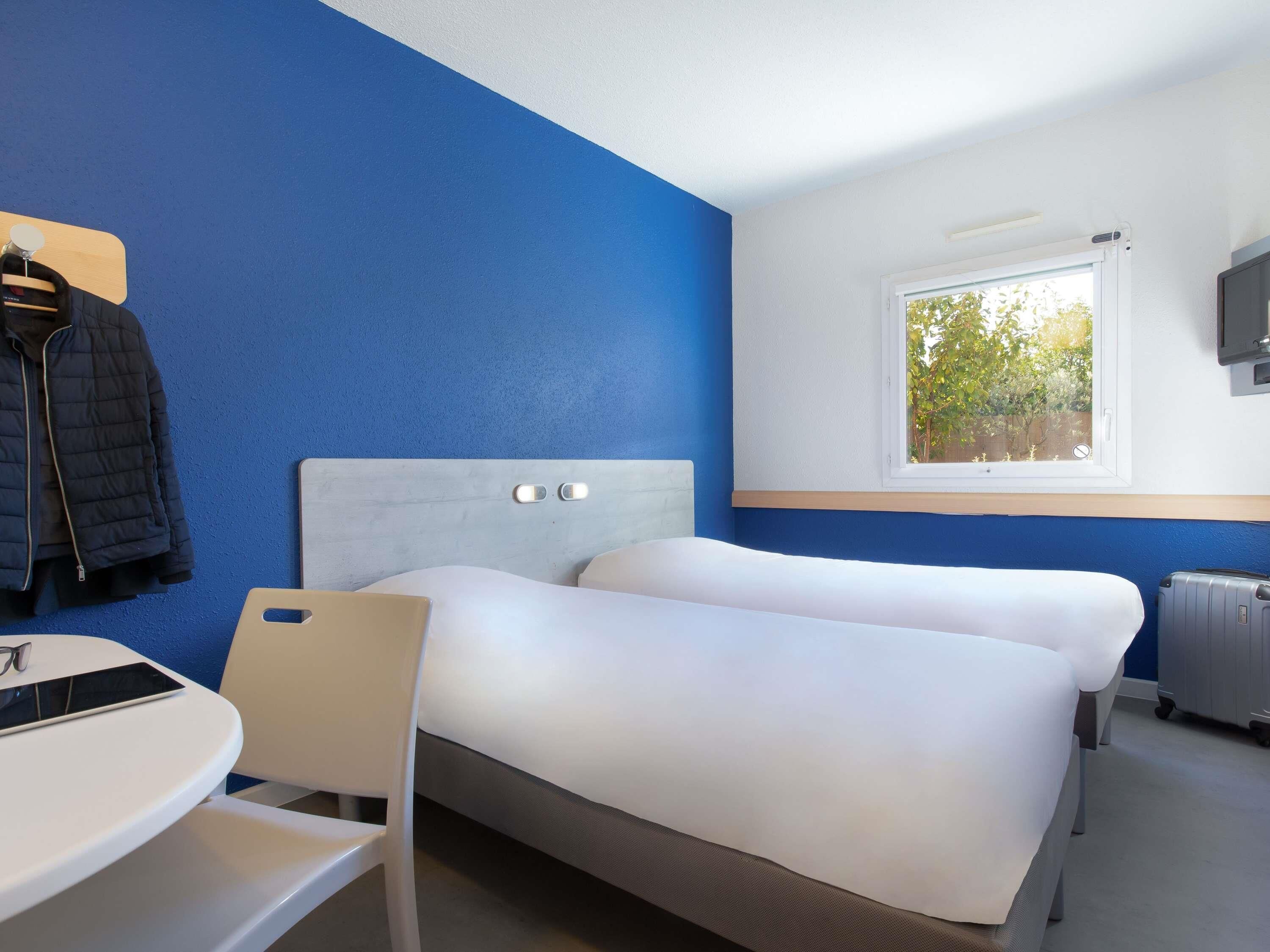 A hotel room with two beds, a desk, a chair, a TV, and a window with a view of trees.