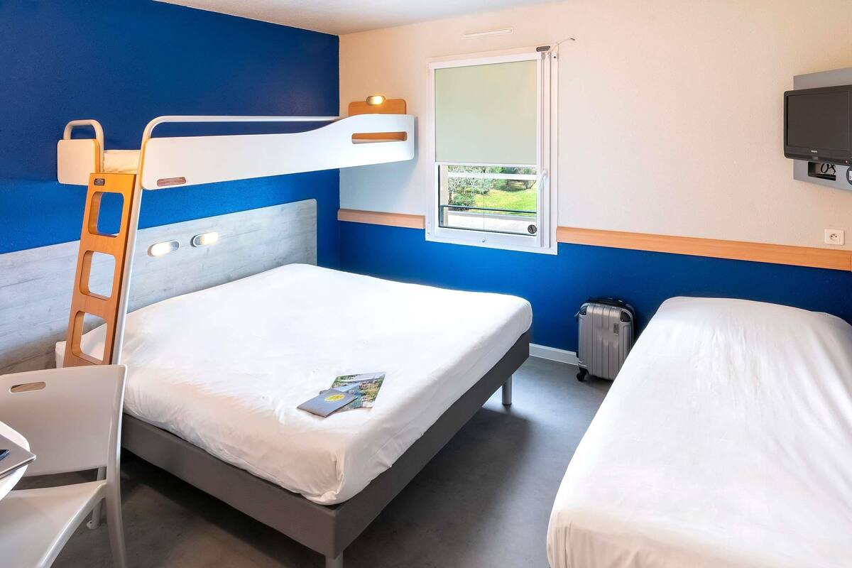 Une chambre d’hôtel avec un lit superposé, un bureau, une chaise, une valise, une fenêtre donnant sur un paysage agréable et un téléviseur fixé au mur.