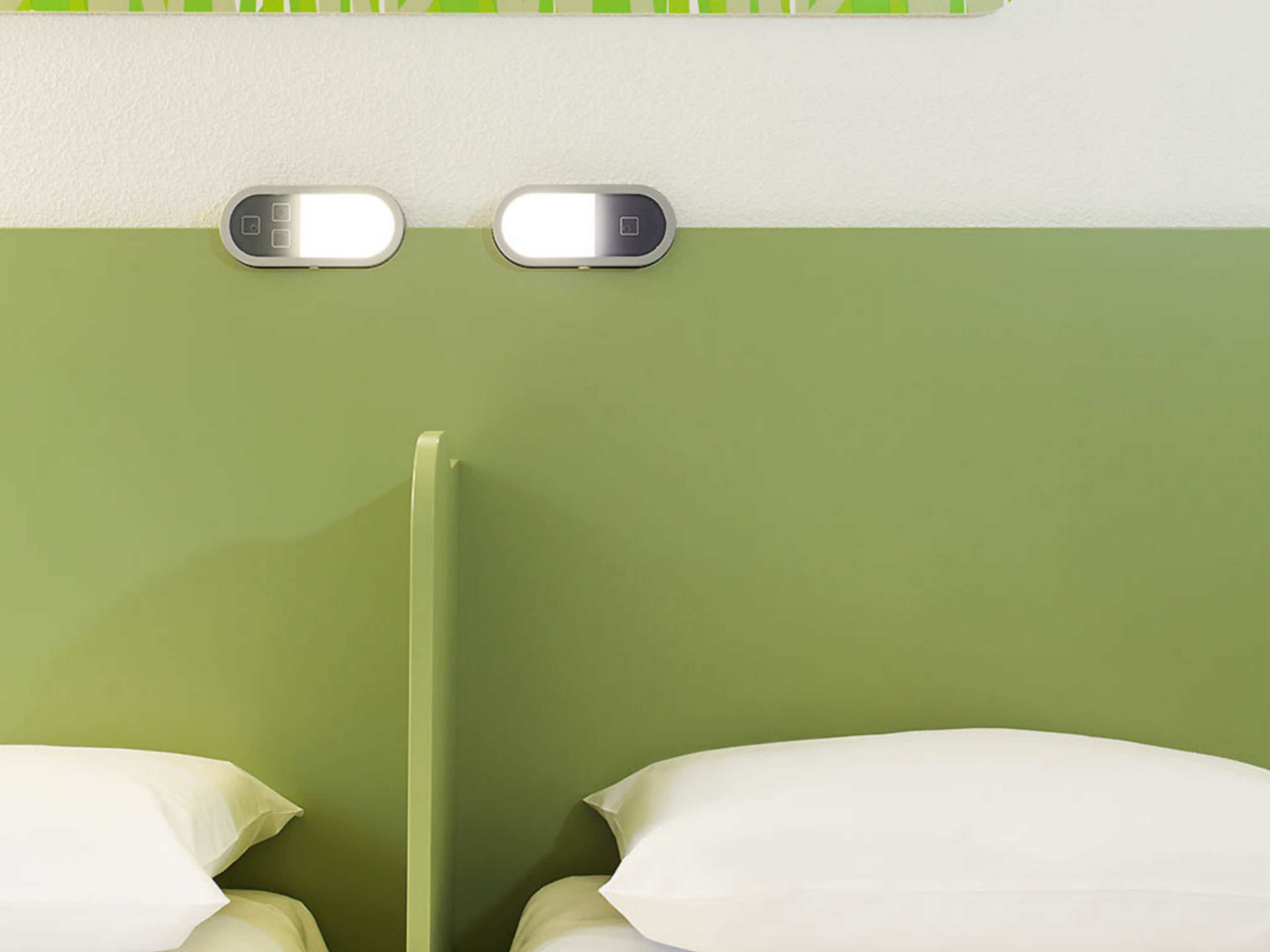 A room with two beds, each with white pillows, and a green wall. There are two illuminated switches on the wall.