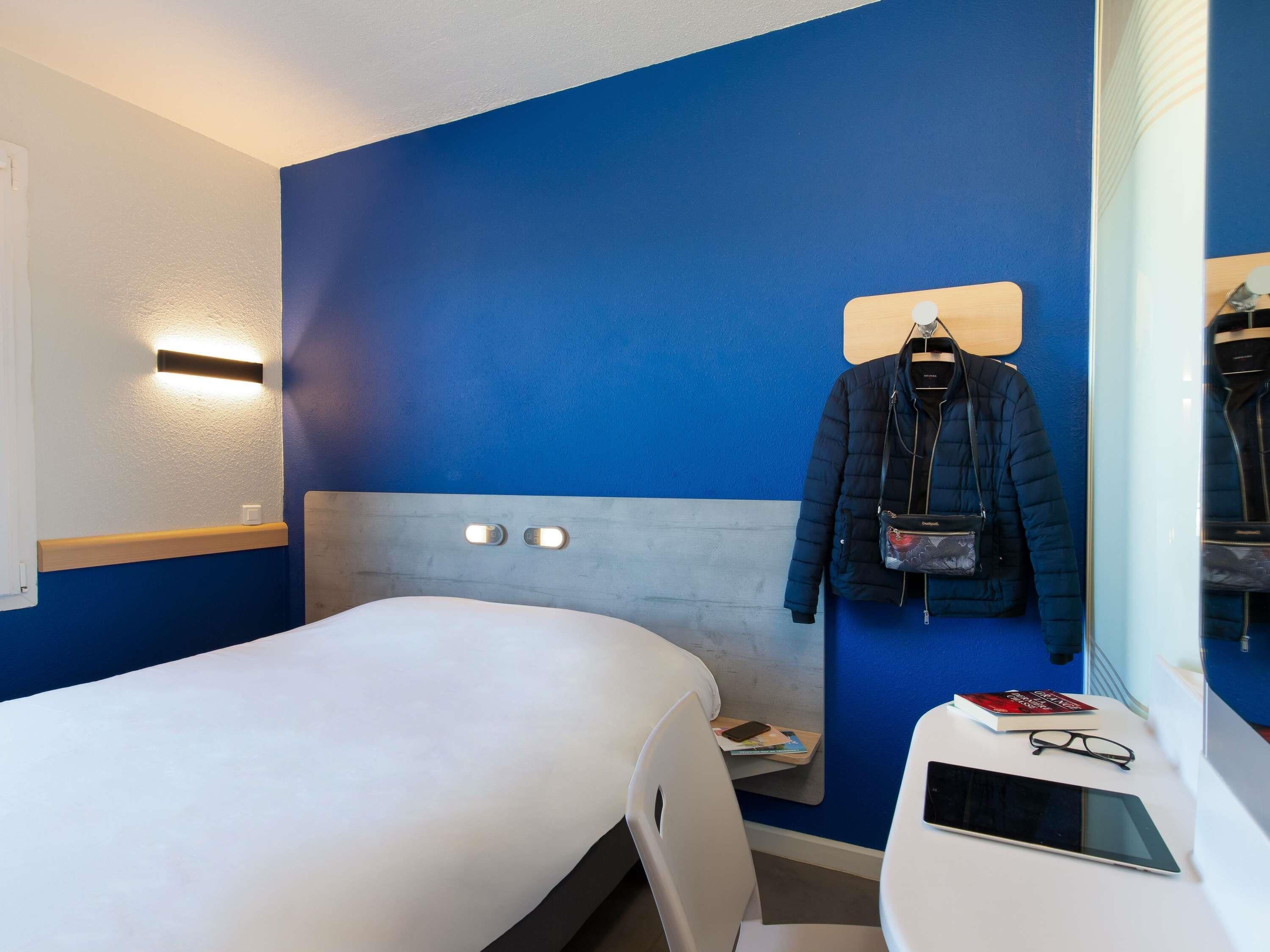 A hotel room with a bed, a desk with a tablet and glasses, a jacket hanging on the wall, and a small shelf with books and a phone.