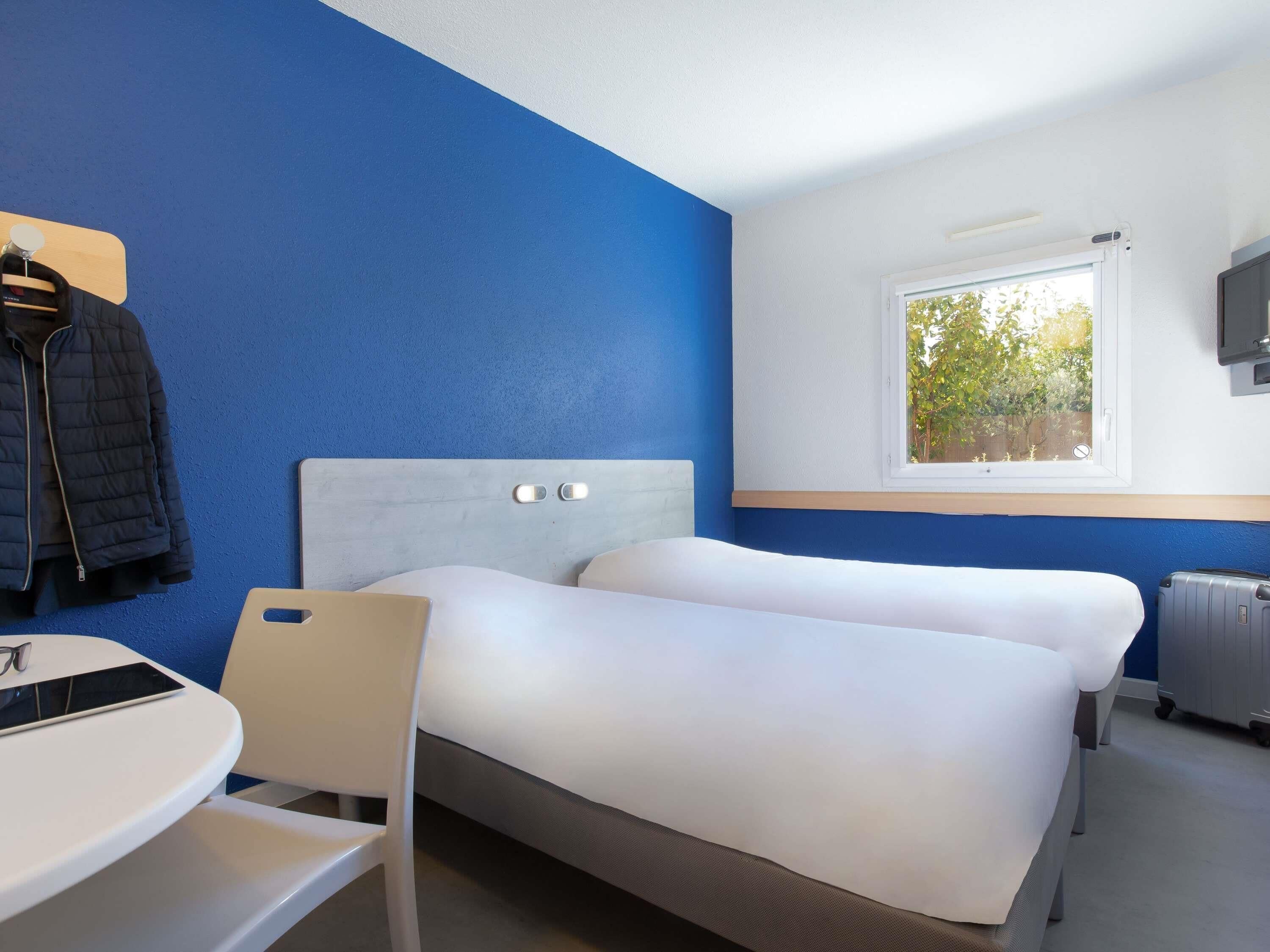 Une chambre d’hôtel avec deux lits, un bureau, une chaise, une télévision et une fenêtre donnant sur des arbres.