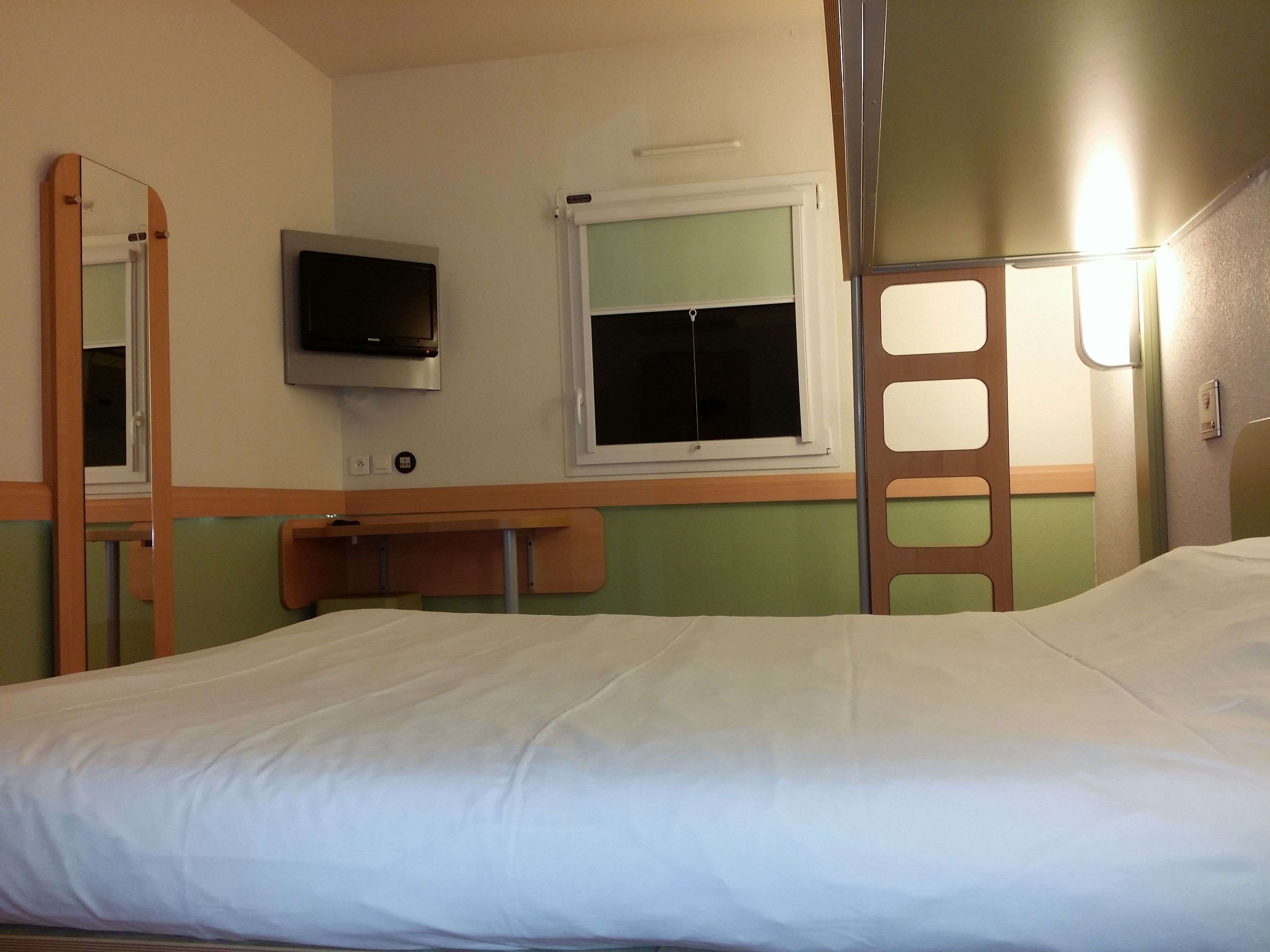 Ein Einzelbett mit weißer Bettwäsche, ein an der Wand montierter Fernseher, ein Fenster mit Jalousien und ein Holztisch.