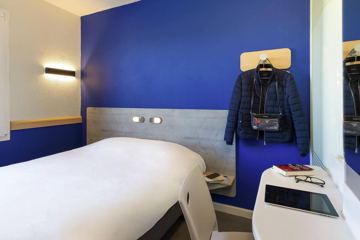 Une chambre d’hôtel avec un lit, un petit bureau avec une chaise, un miroir et un porte-manteau fixé au mur, sur lequel sont accrochés un manteau et un sac.