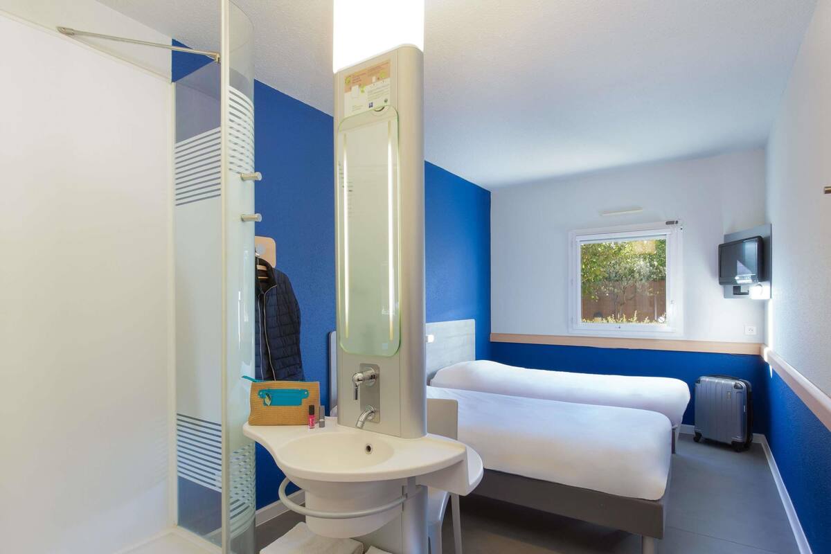A compact hotel room with a bed, a sink, a mirror, a window, and a TV.