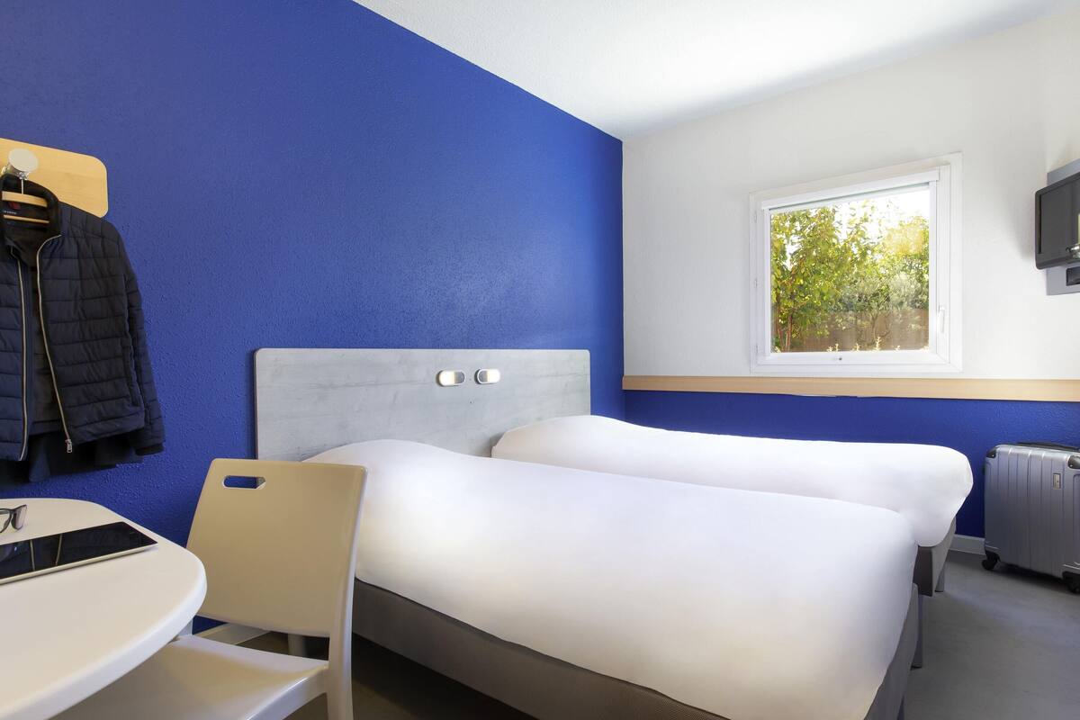 Une chambre d’hôtel avec deux lits, un bureau, une chaise, un porte-manteau et une fenêtre donnant sur des arbres.