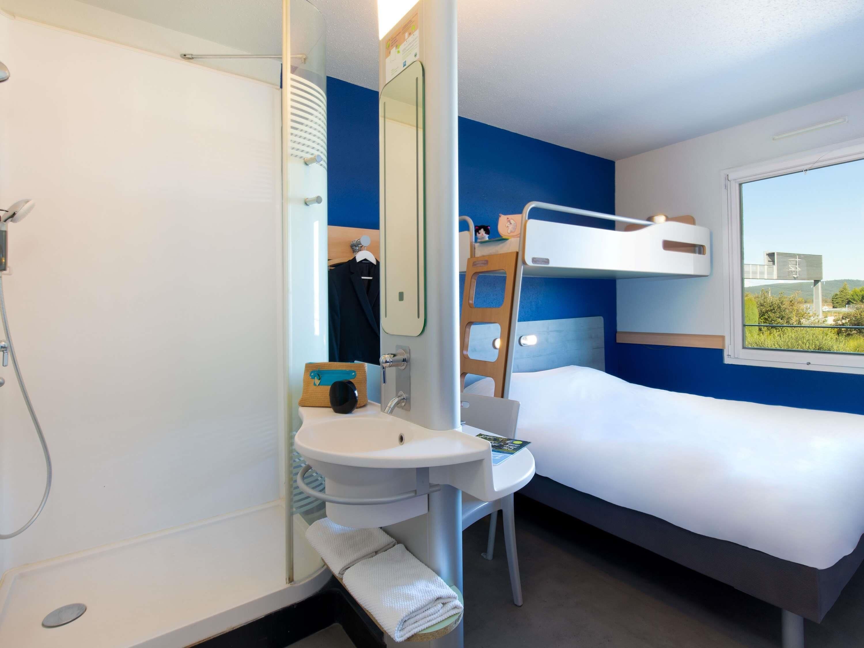A compact hotel room with a bunk bed, a small bathroom with a sink and mirror, and a window offering a view of greenery.