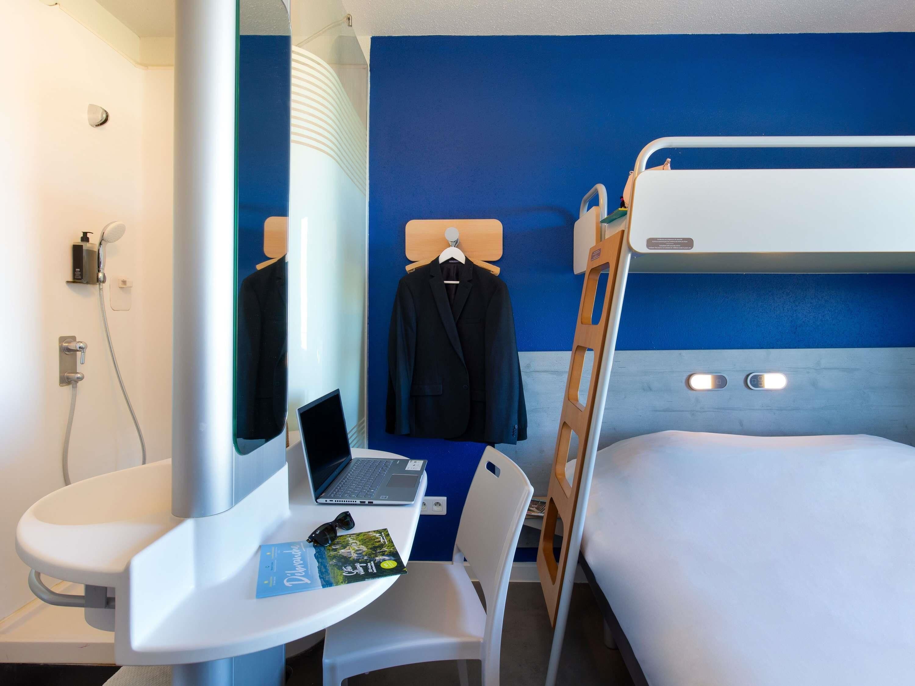 A hotel room with a bunk bed, a desk with a laptop, a mirror, a towel rack with a coat, and a small sink.
