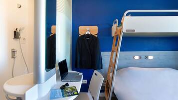 Habitación de hotel con literas, un escritorio con una computadora portátil, un espejo, un perchero con abrigo y un lavamanos pequeño.