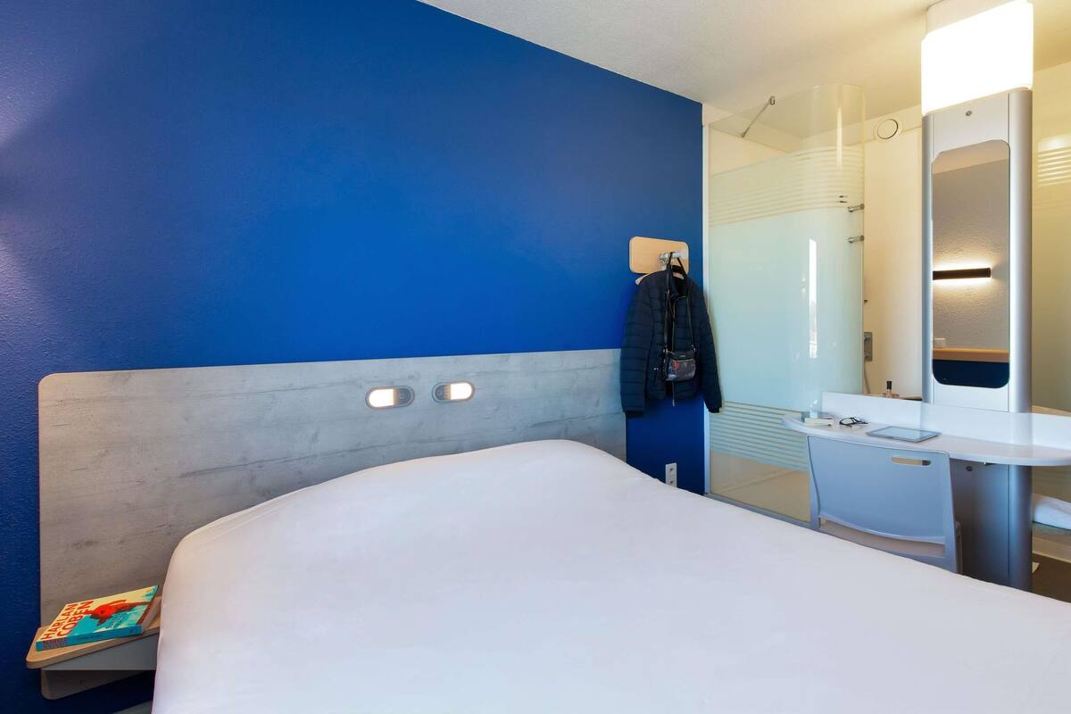 A hotel room with a large bed, a bedside table with a book, a mirror, and a bathroom visible through a glass partition.