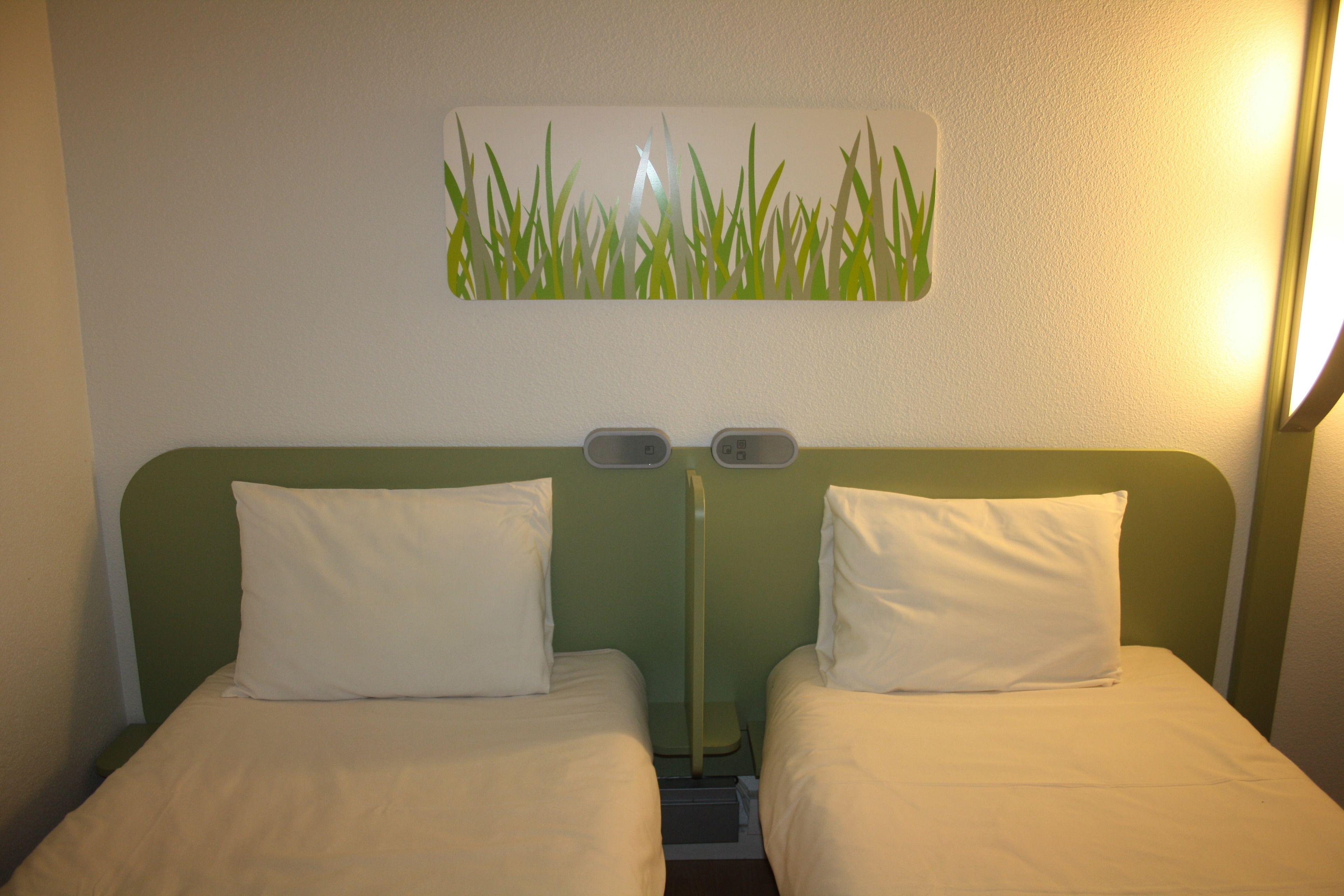 Ett hotellrum med två separata sängar, en grön sänggavel och ett väggmonterat konstverk som föreställer gräs.