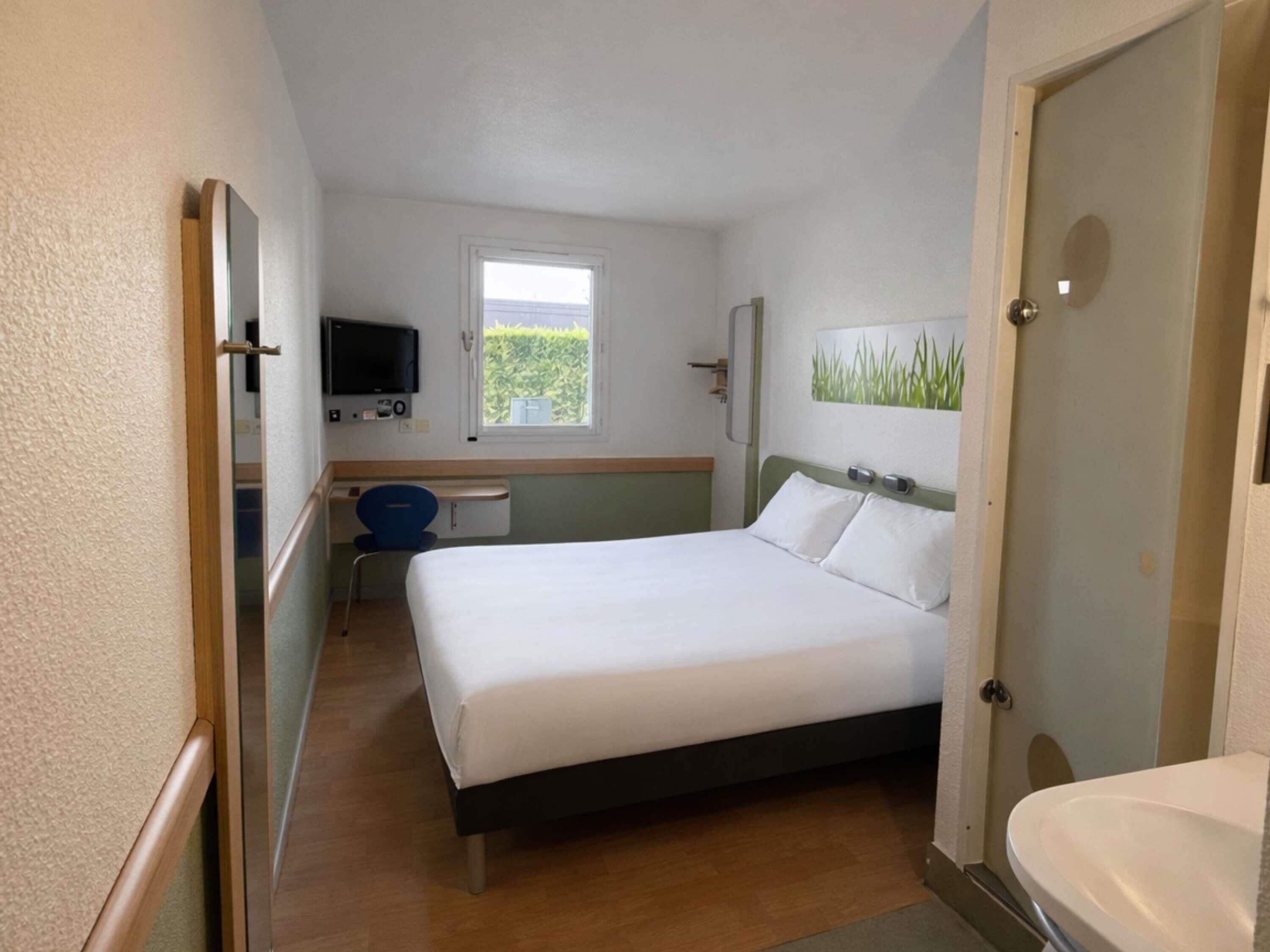 Quarto de hotel com cama, escrivaninha, televisão e janela.