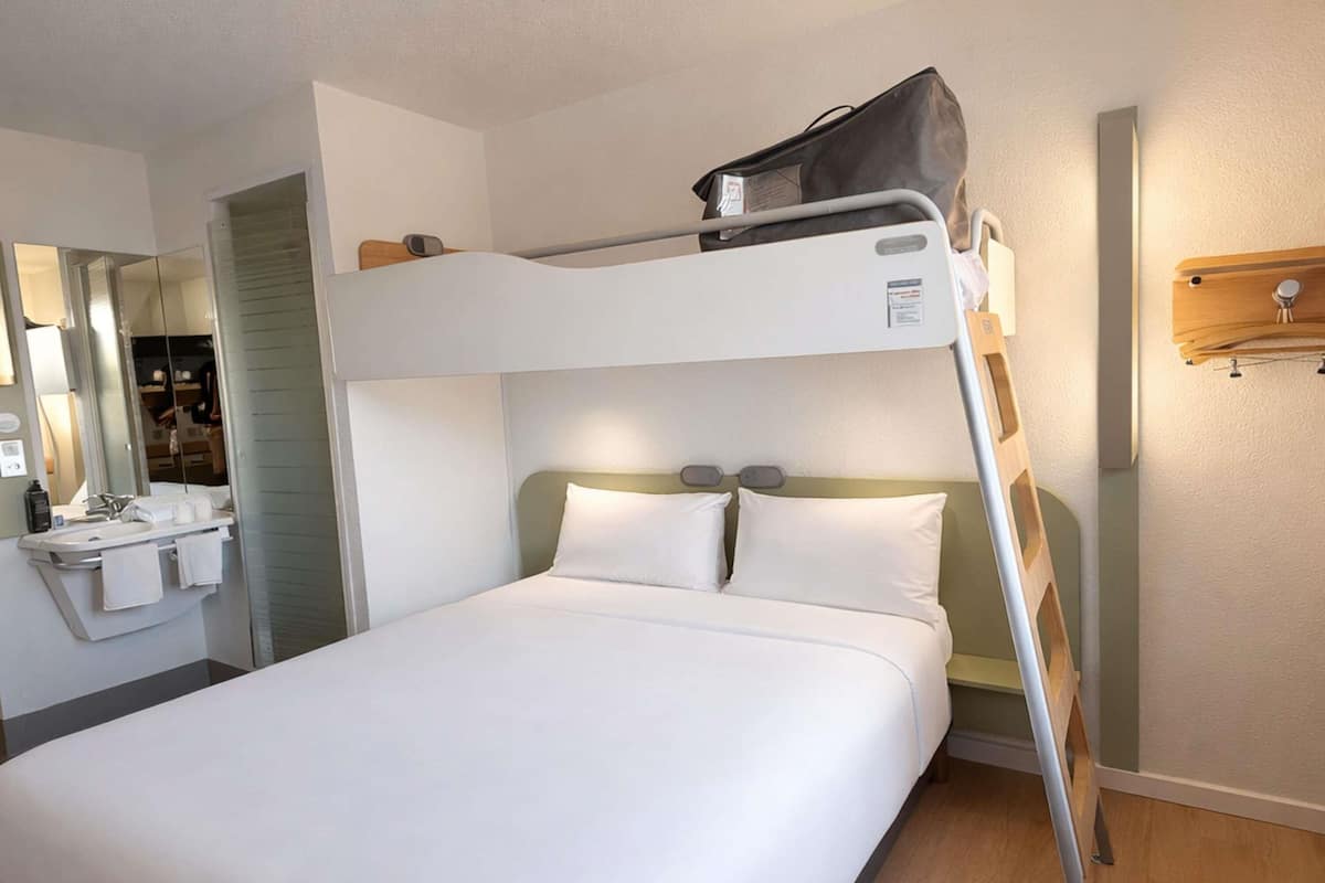 Una cama individual superpuesta con una maleta encima, una mesita de noche con una lámpara y un espejo.