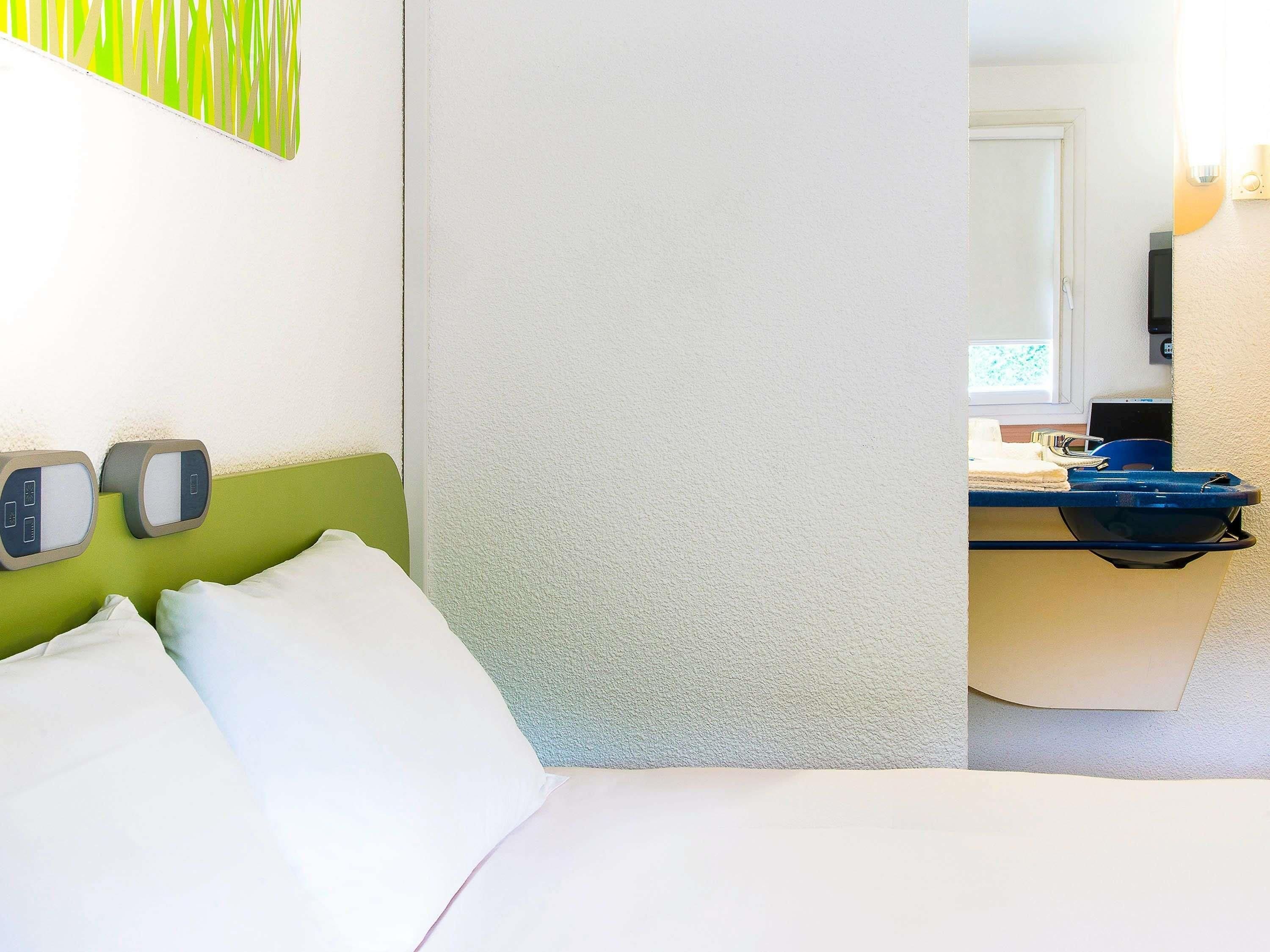 Habitación de hotel con cama, cabecera verde, televisor montado en la pared y baño visible a través de una puerta abierta.