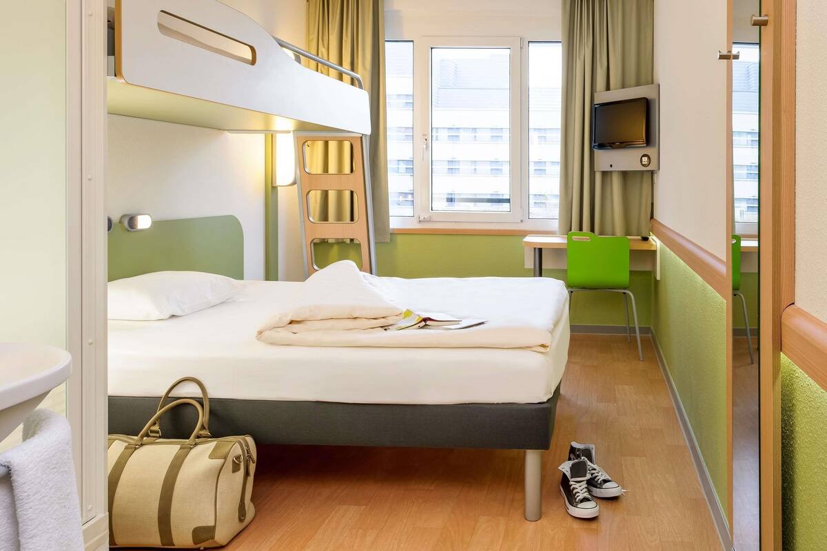 A hotel room with a bunk bed, a desk, a chair, a TV, and a window with curtains.
