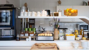 A kitchen counter with a coffee machine, jars of condiments, a bread loaf, and a basket of oranges. - ibis budget Montauban Les Chaumes (Montauban)