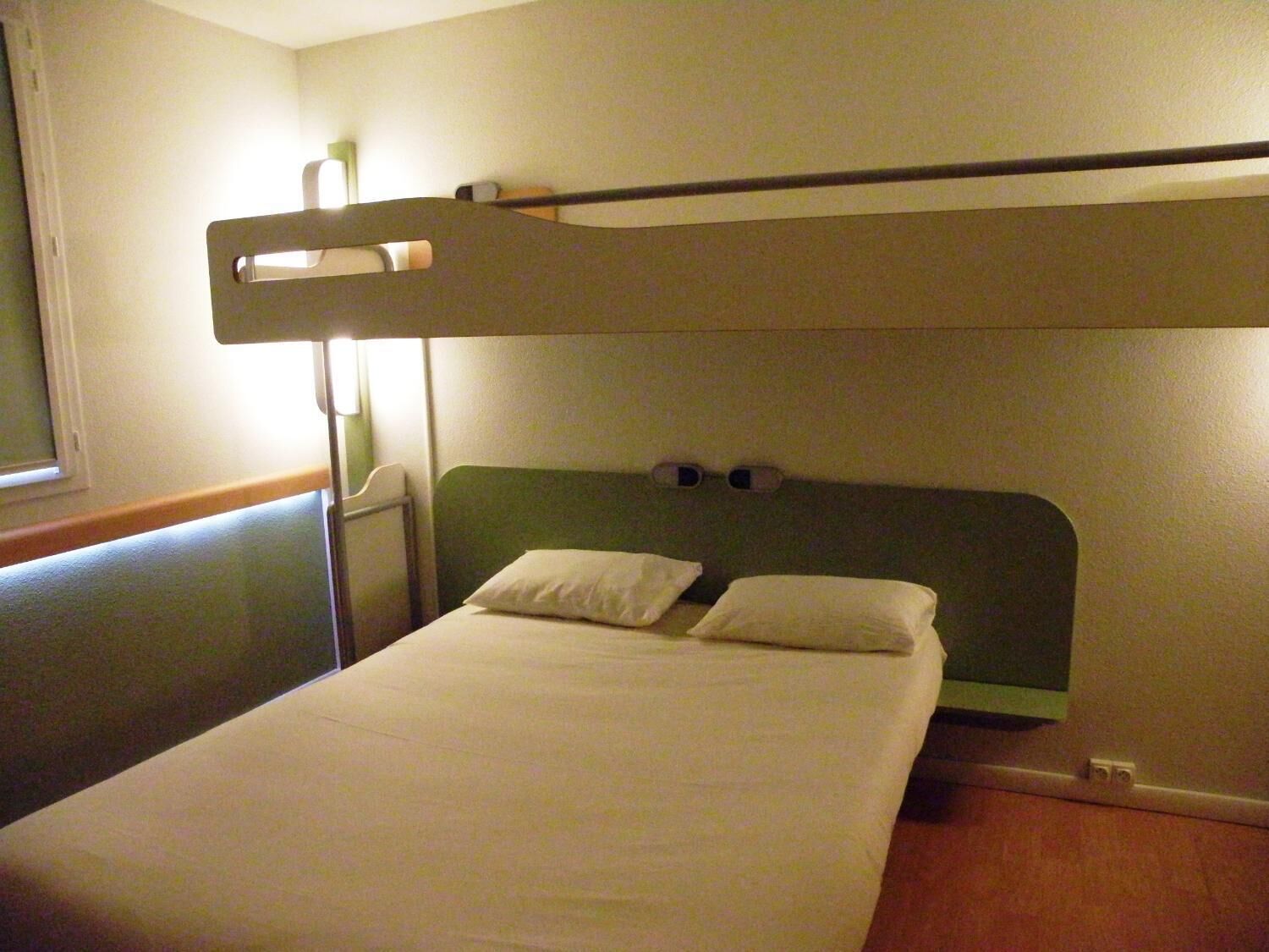 Een stapelbed met een groen hoofdeinde, wit beddengoed en een nachtkastje met een lamp.