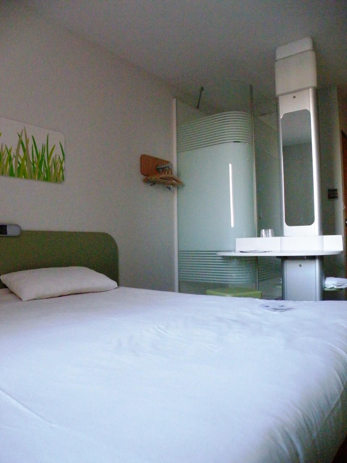 A hotel room with a bed, a small bedside table, a mirror, and a wall-mounted shelf.