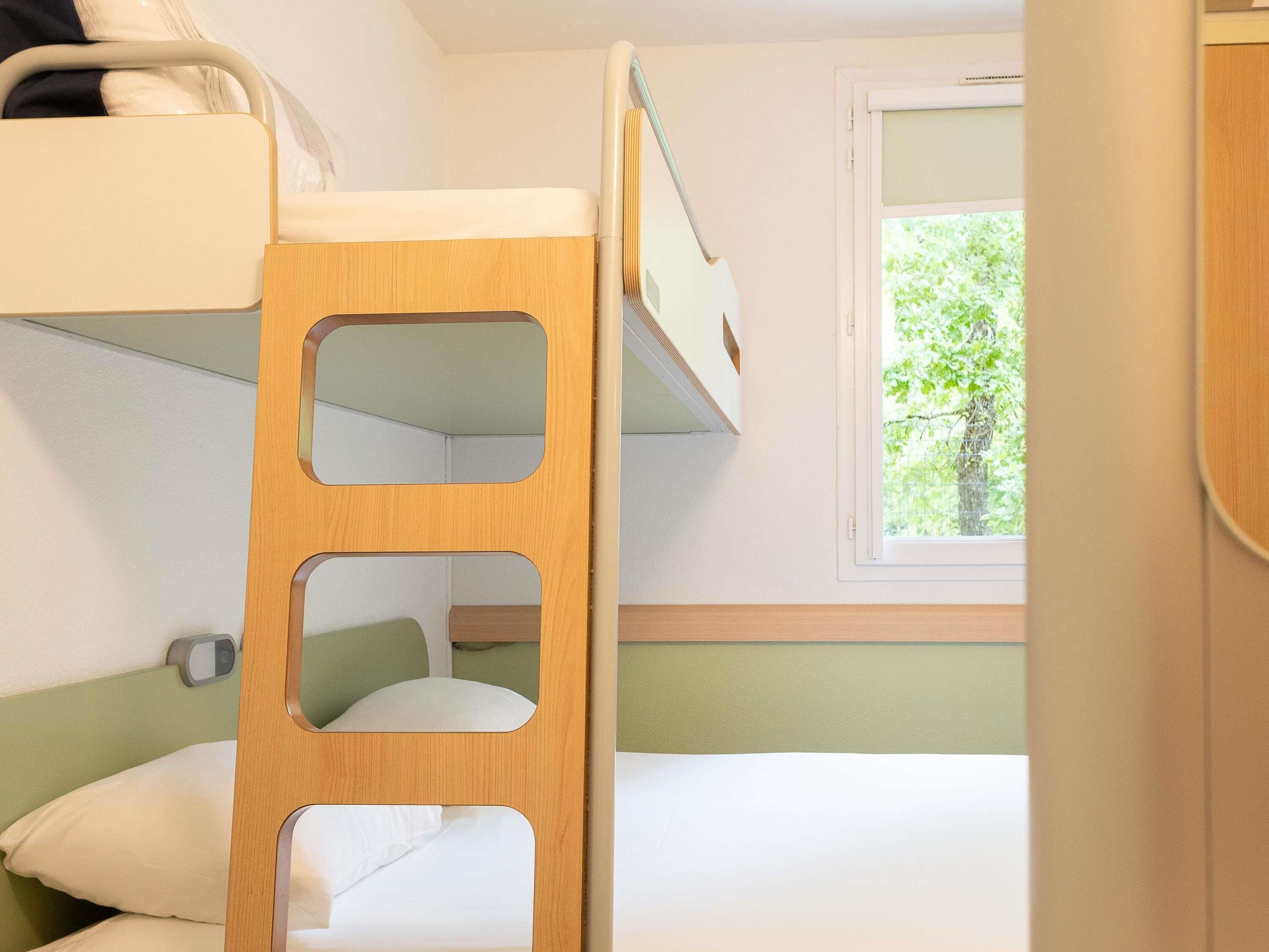 A bunk bed with a wooden ladder, a bedside table, and a window with a view of greenery.
