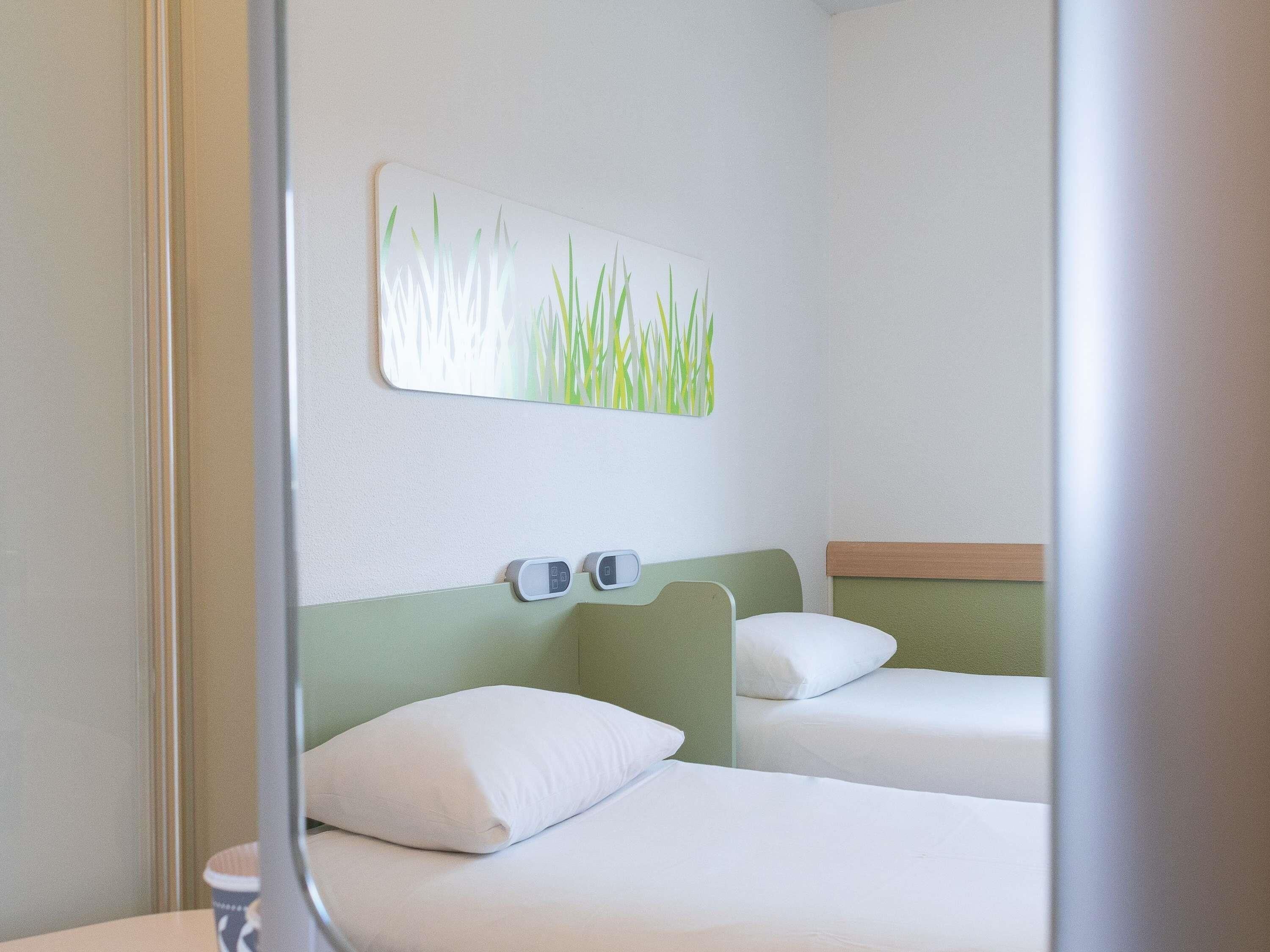 Une chambre d’hôtel avec deux lits, un interrupteur fixé au mur et un tableau représentant de l’herbe.