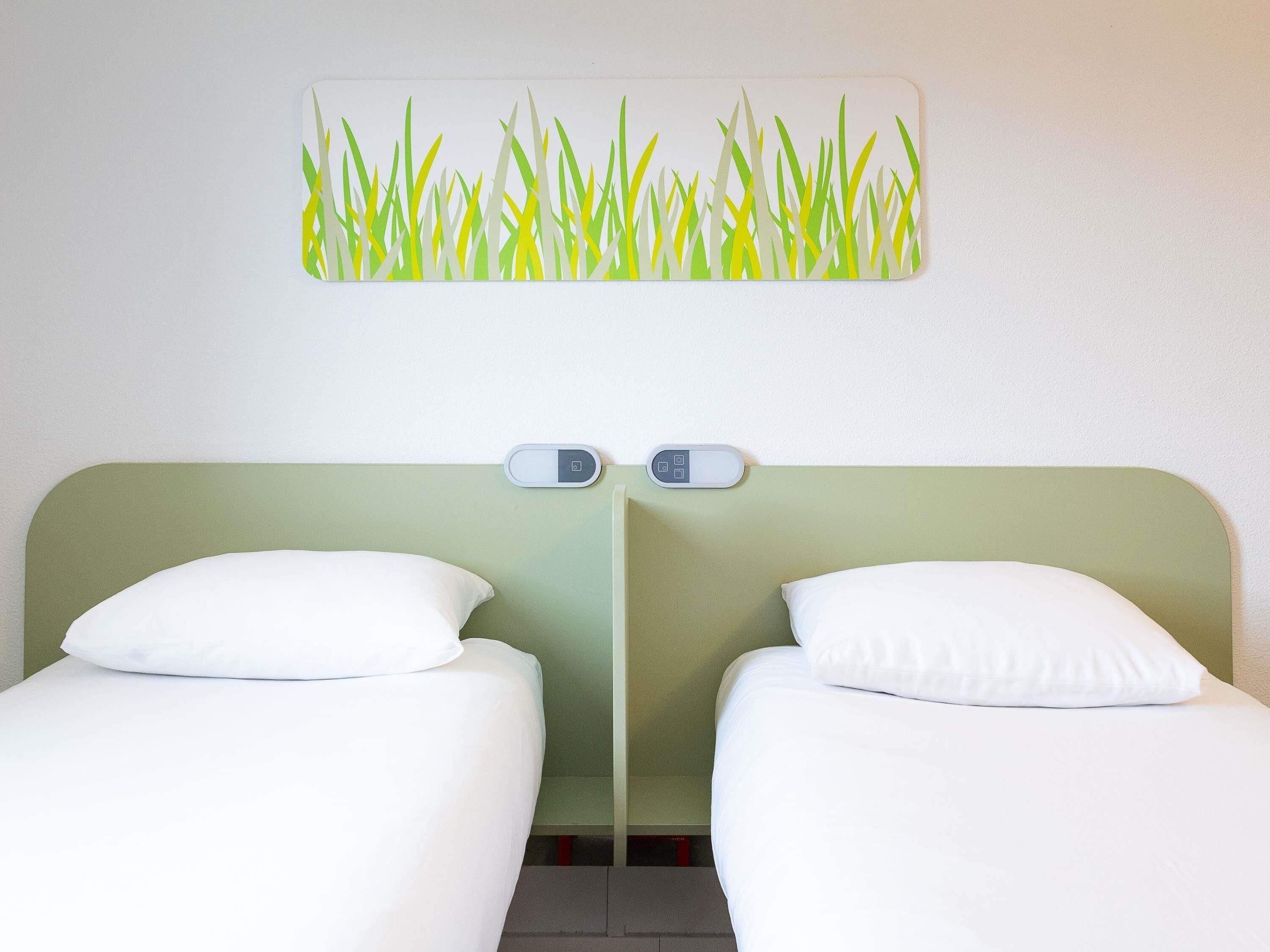 A hotel room with two single beds, a wall-mounted light switch, and a painting of grass above the beds.