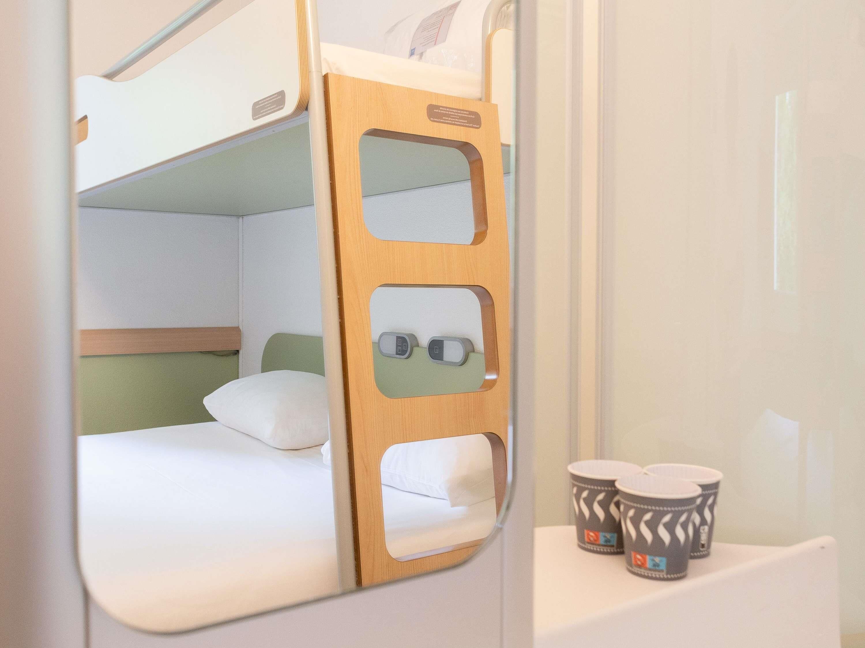 Cama litera con cabecera de madera, un pequeño estante con dos tazas estampadas y un espejo que refleja la habitación.