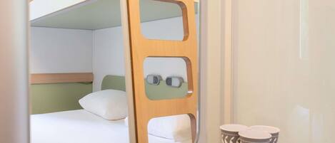 Cama litera con cabecera de madera, un pequeño estante con dos tazas estampadas y un espejo que refleja la habitación.