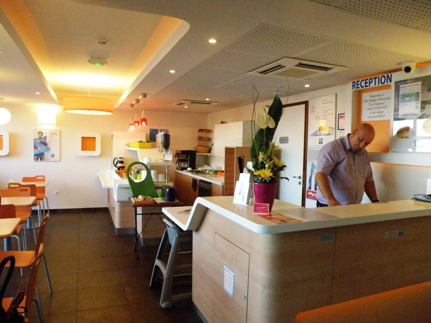 A reception area with a counter, chairs, tables, and a man standing behind the counter.