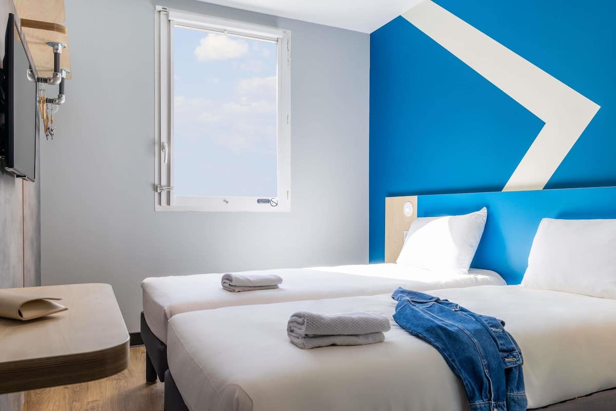 A hotel room with two beds, a window, a desk, and a blue accent wall.