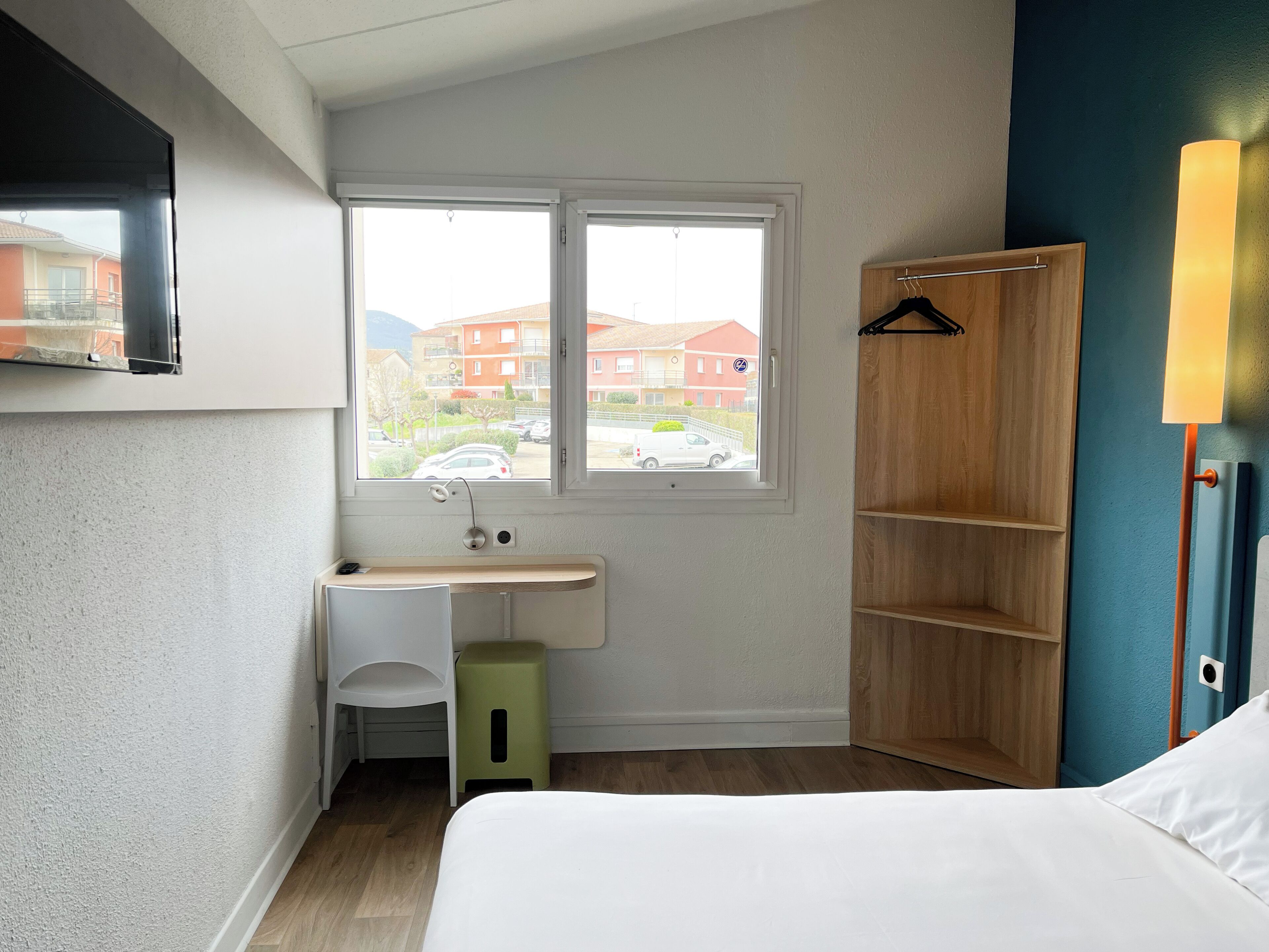 Ein kleiner, gut beleuchteter Raum mit einem Bett, einem Schreibtisch mit Stuhl, einem Fenster mit Blick auf Gebäude und einem Holzschrank.