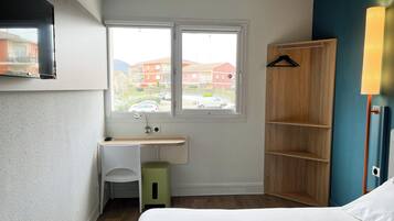 Ein kleiner, gut beleuchteter Raum mit einem Bett, einem Schreibtisch mit Stuhl, einem Fenster mit Blick auf Gebäude und einem Holzschrank.