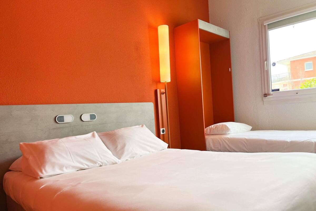 Habitación de hotel con dos camas, una mesita de noche, una lámpara de pared y una ventana con vistas a edificios.