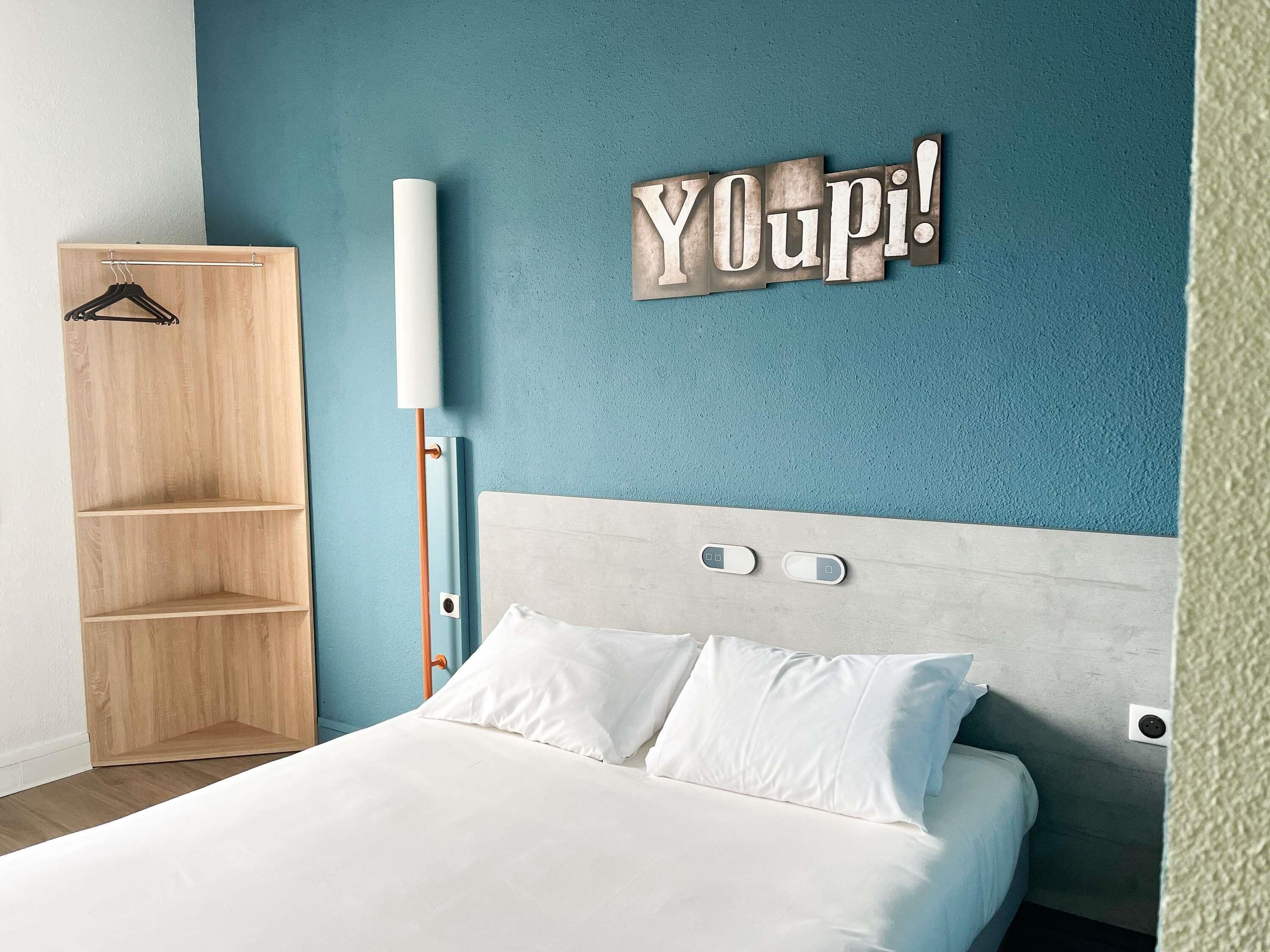 A bedroom with a bed, a wooden wardrobe, a wall-mounted light, and a 'YOUPI!' sign.