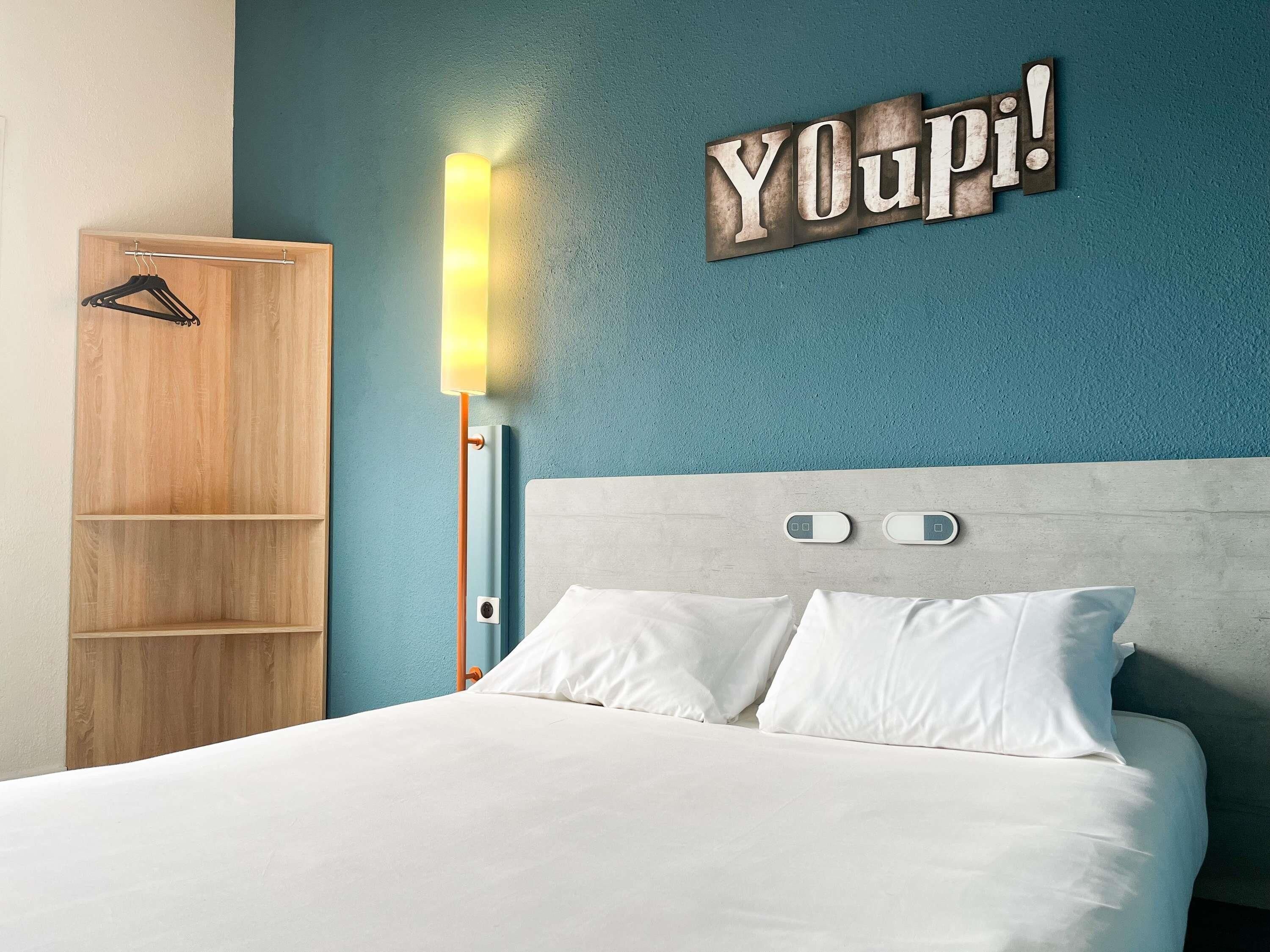 A bedroom with a bed, a wooden wardrobe, a wall-mounted light, and a 'YOUpi!' sign.