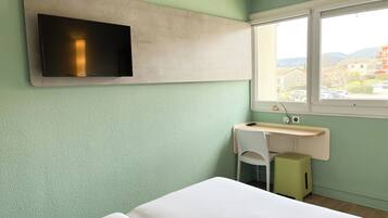 Una habitación con cama, escritorio, silla, televisor montado en la pared y ventana con vista a edificios.