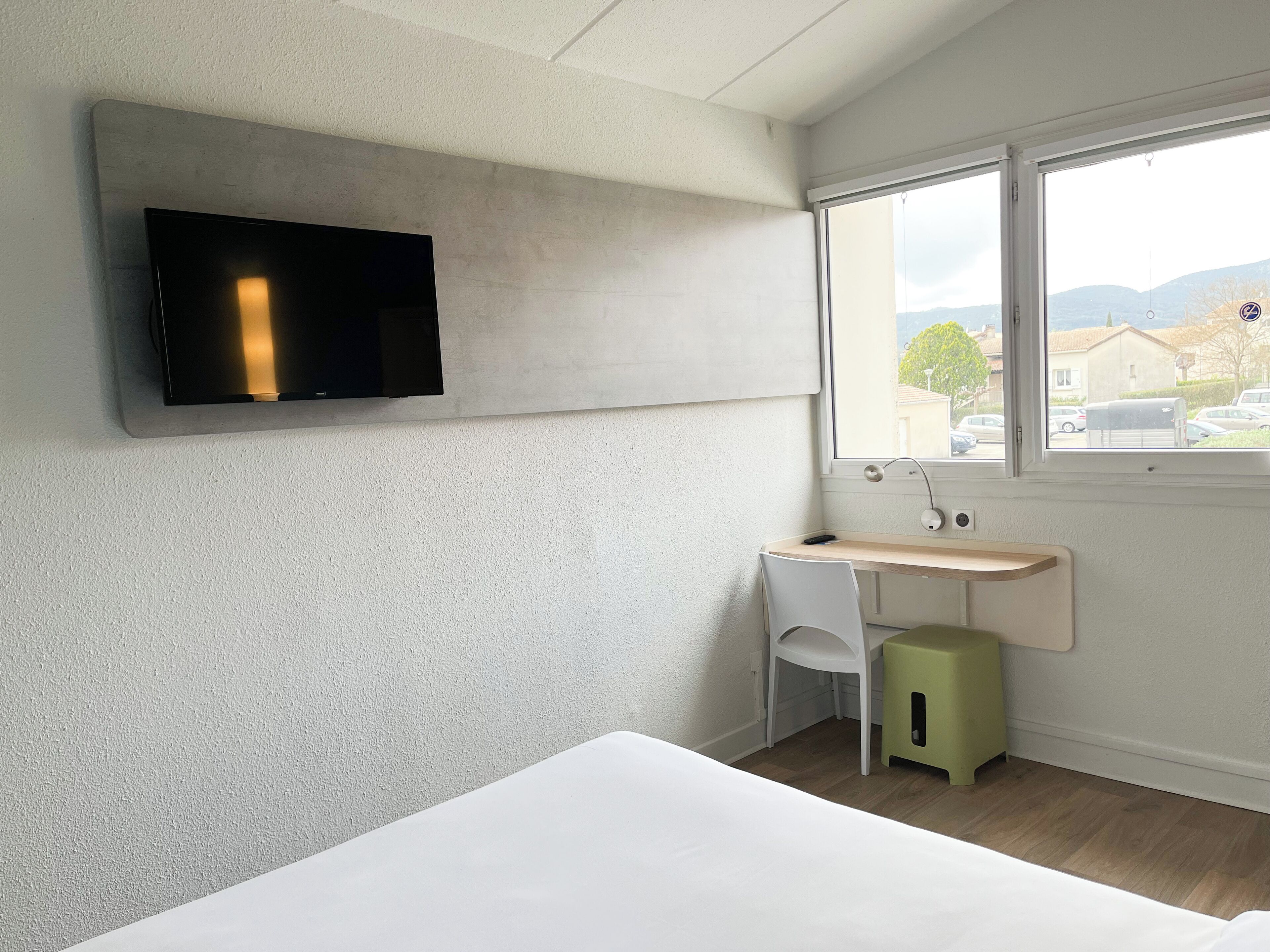 Une chambre d’hôtel moderne équipée d’un lit, d’un bureau avec une chaise, d’un téléviseur fixé au mur et d’une fenêtre donnant sur l’extérieur.