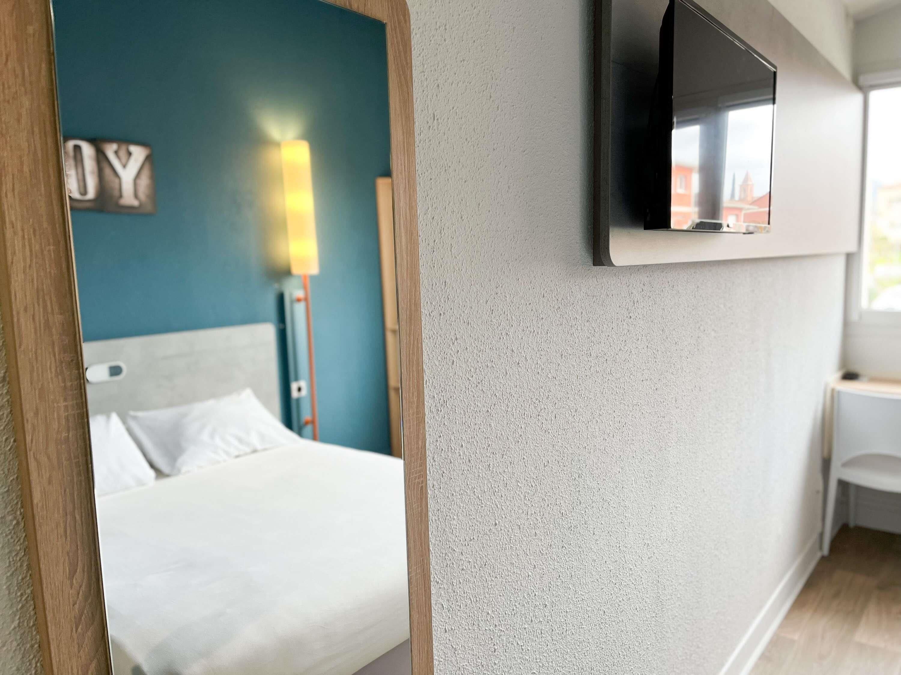 Hotellihuone, jossa on sänky, seinälle asennettu televisio ja seinävalaisin.