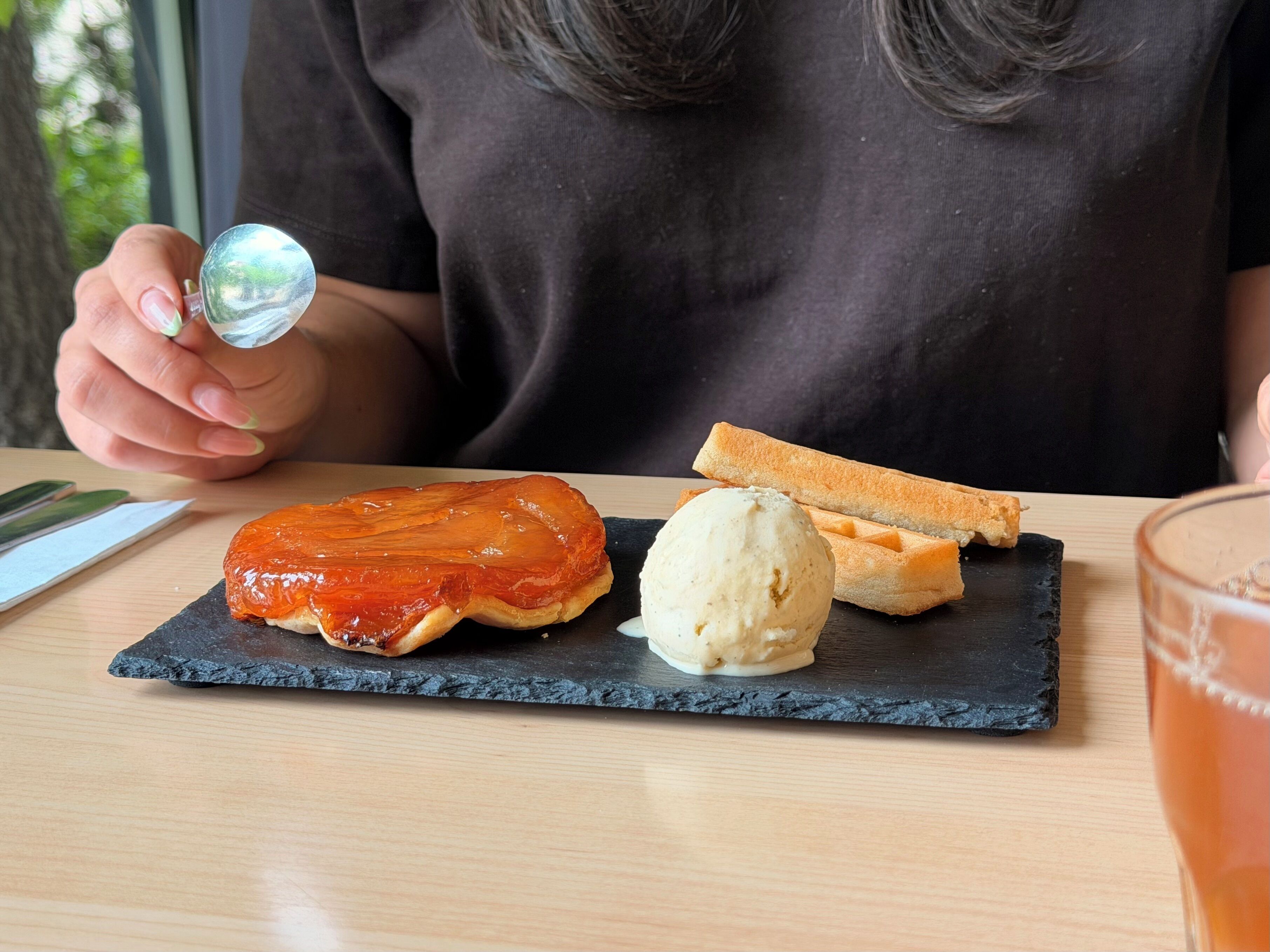Una persona sosteniendo un vaso, un plato con un pastel glaseado y una porción de helado, y un trozo de pan sobre un plato de pizarra oscura.