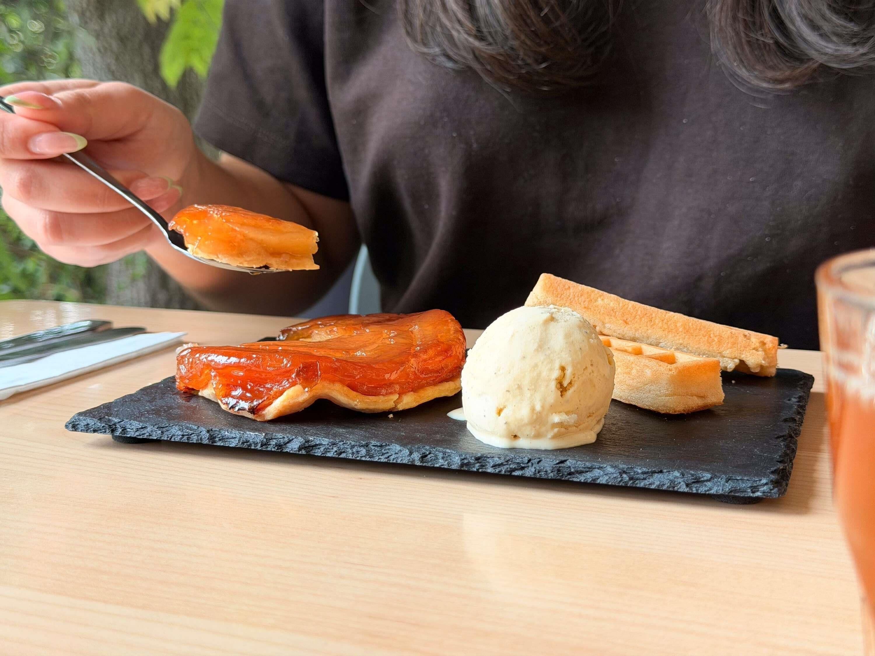 Eine Person hält einen Löffel mit einer Portion Speise, einem runden Dessert und zwei Brotscheiben auf einem Schieferteller.