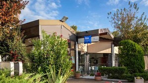 A modern hotel with a glass facade, outdoor seating, and a well-maintained garden. - ibis budget Alès Saint Christol (Saint-Christol-Les-Ales)