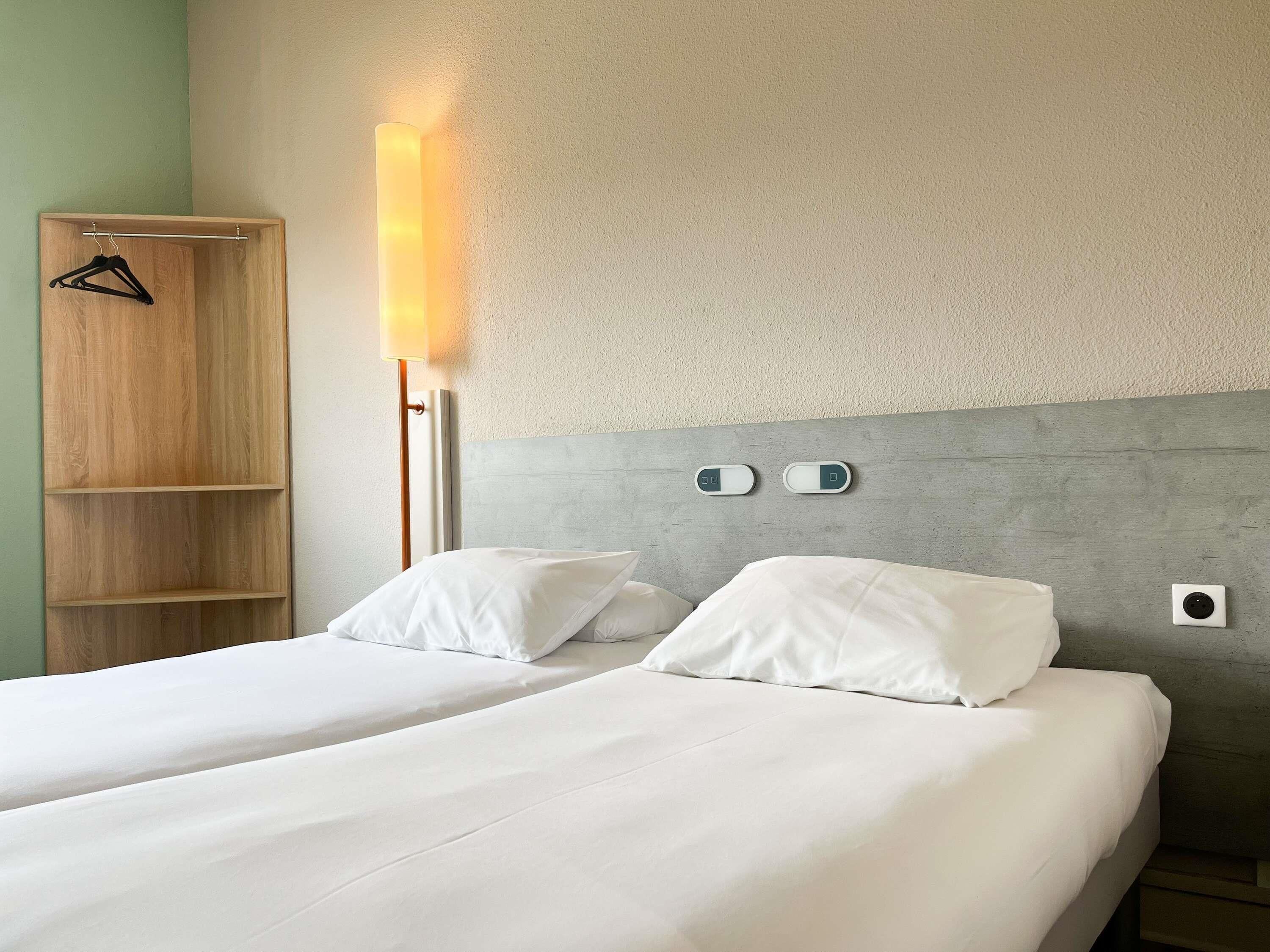 Ein Hotelzimmer mit zwei Betten, einem Holzschrank, einer Nachttischlampe und einer wandmontierten Steuerung.