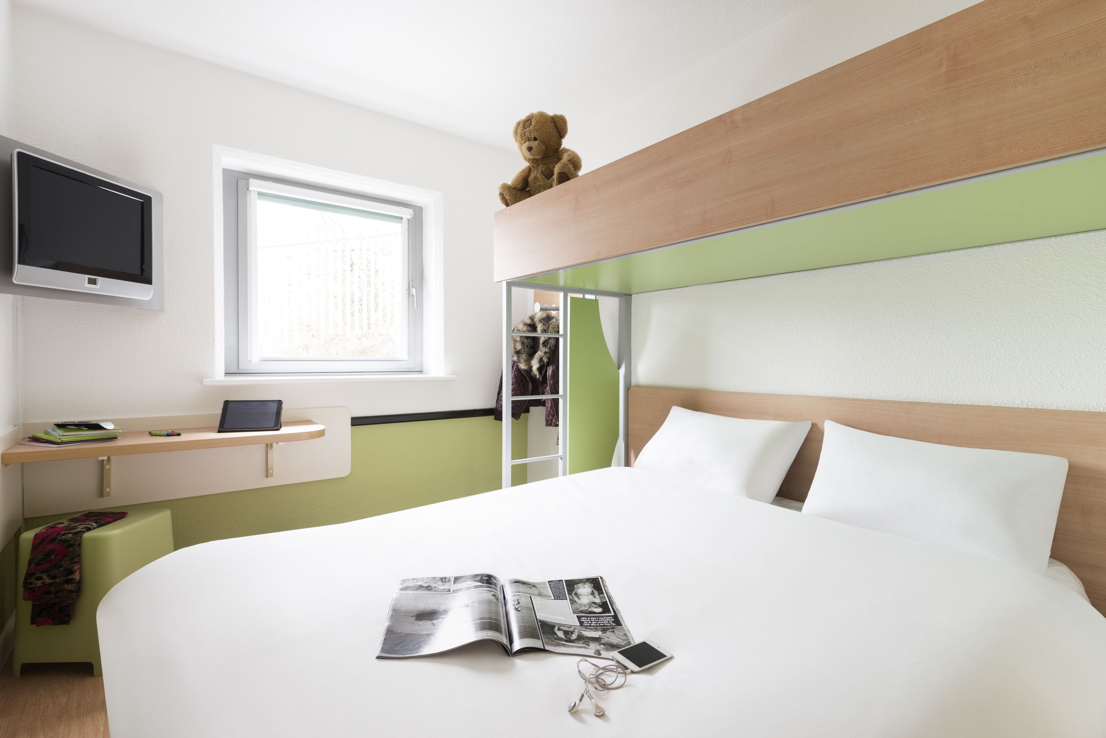 Une chambre d’hôtel moderne avec un grand lit, une table de chevet sur laquelle se trouve un ordinateur portable, un téléviseur fixé au mur et un ours en peluche posé sur la tête de lit.