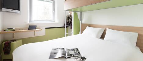 A modern hotel room with a large bed, a bedside table with a laptop, a TV mounted on the wall, and a teddy bear on the bed's headboard.