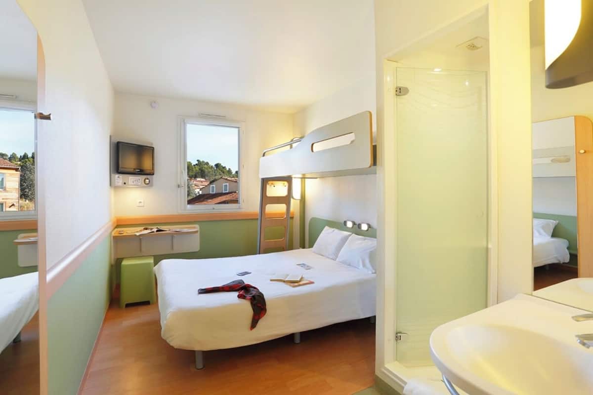 A compact hotel room with a single bed, a small bedside table, a mirror, a television, and a bathroom with a sink and shower.