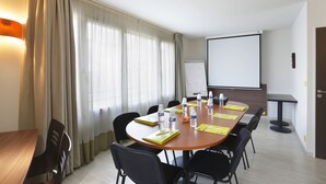 A conference room with a wooden table, black chairs, a whiteboard, and a projector screen. - First Inn Hotel (Apt)