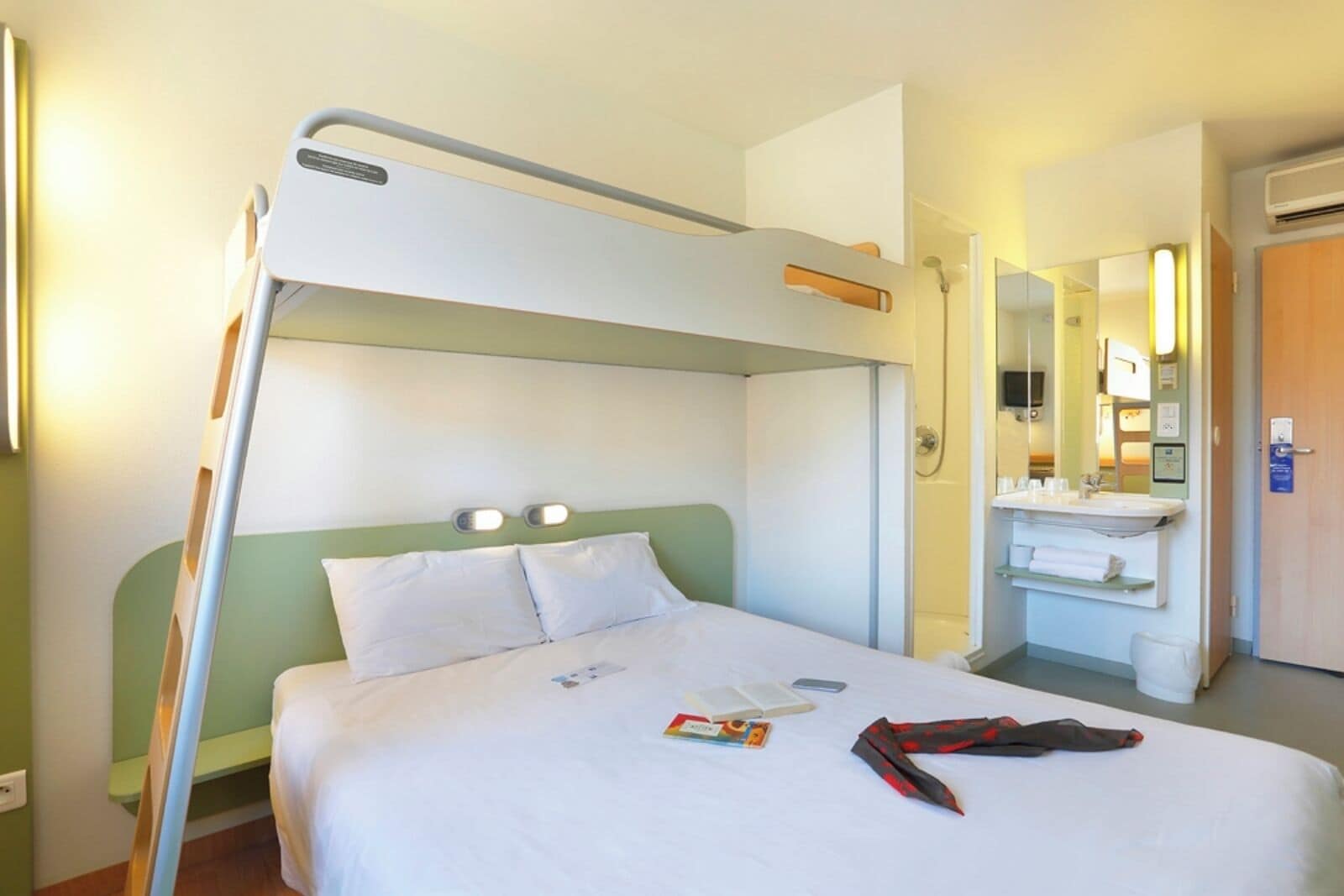 A hotel room with a bunk bed, a bedside table with a book and a phone, a bathroom with a sink and mirror, and a door with a sign.