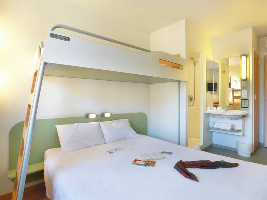 Une chambre d’hôtel avec un lit superposé, une table de chevet avec un livre et un téléphone, une salle de bain avec un lavabo et un miroir, et une porte avec une pancarte.